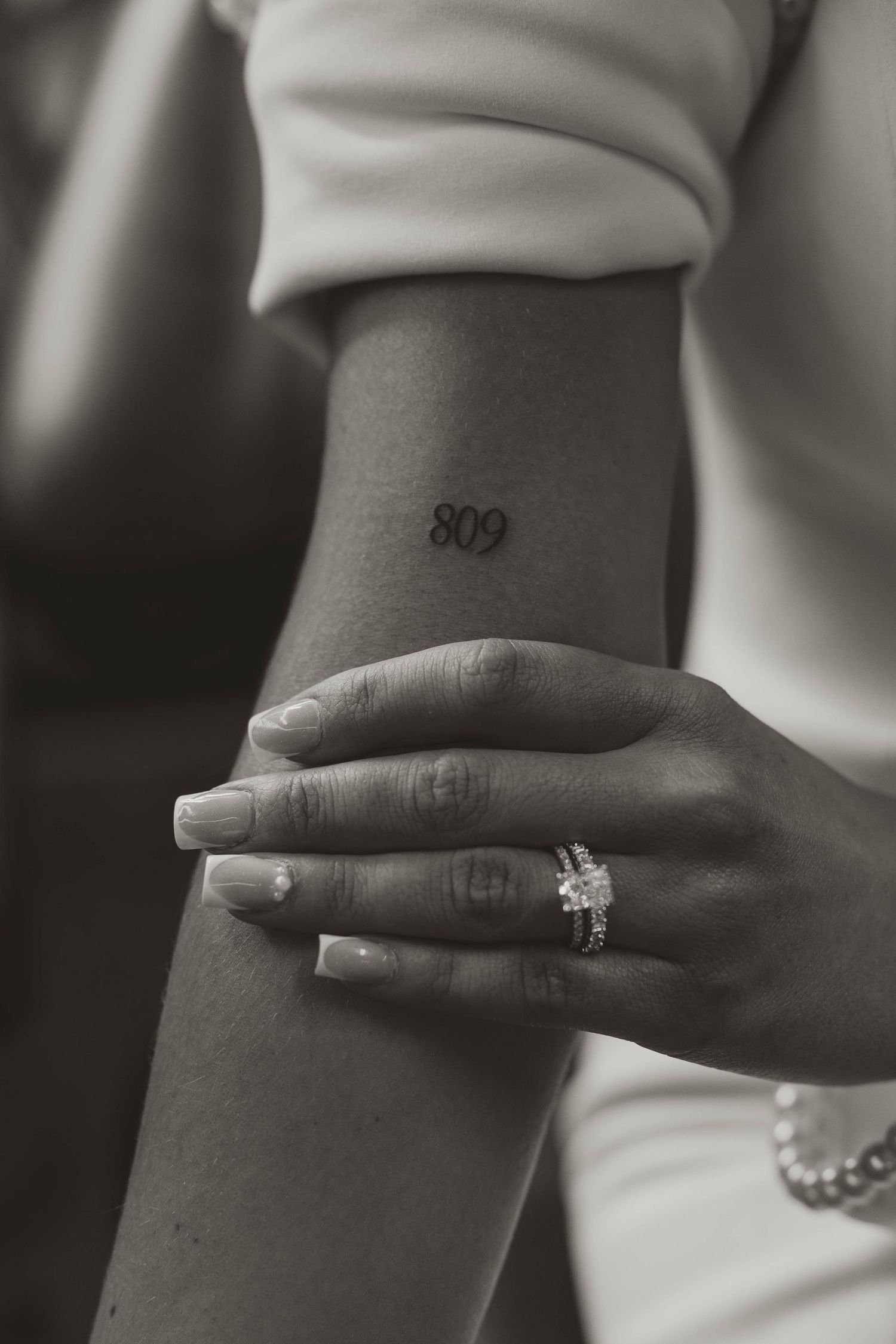 Black and white photo of a woman's arm with an 809 tattoo on the wrist, wearing a diamond ring and pearl bracelet.