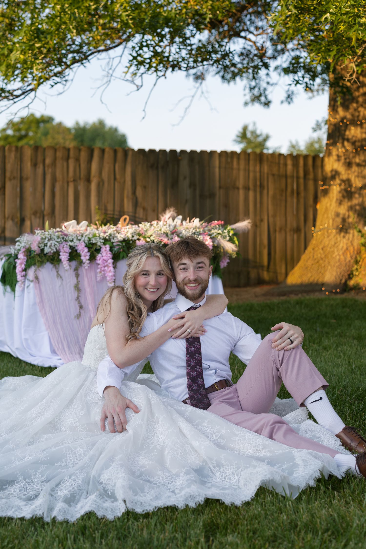 A smiling bride and groom sit together on green grass at an outdoor wedding with a floral-decorated table behind them.