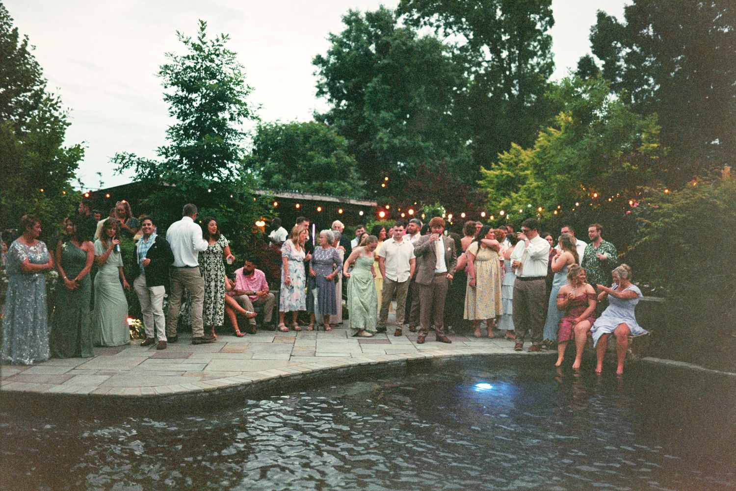 Outdoor poolside party with guests socializing under string lights surrounded by lush green trees at dusk.