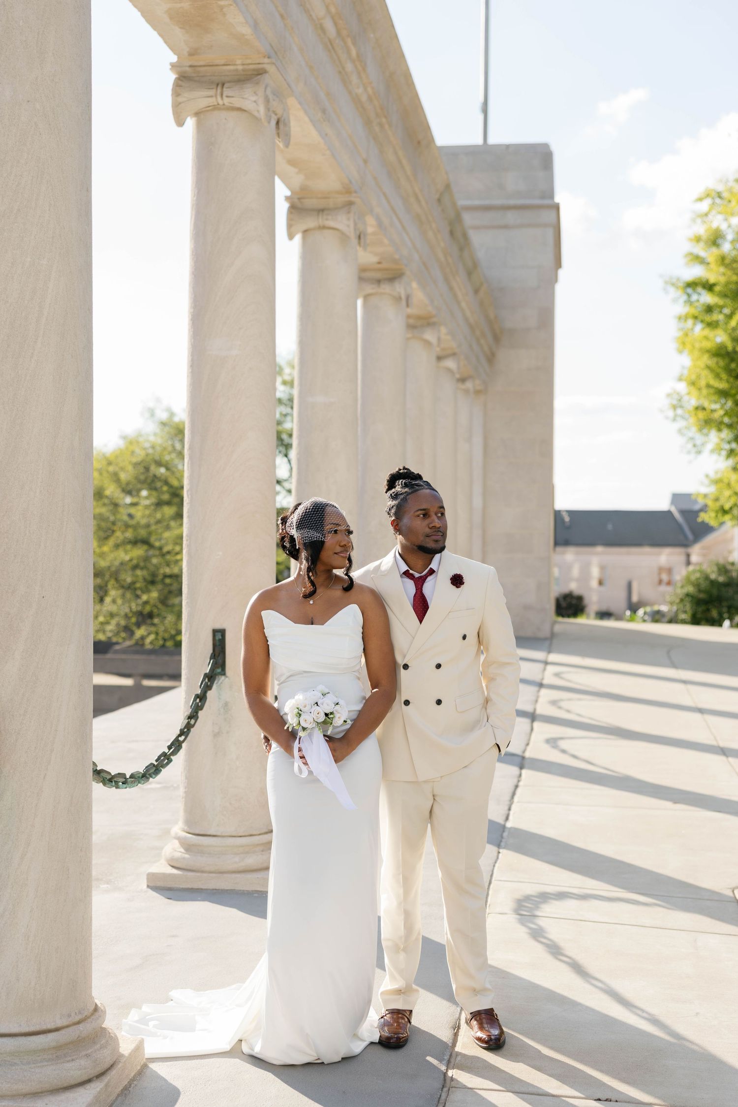 Bride and groom in cream wedding attire pose elegantly among white marble columns on their wedding day.