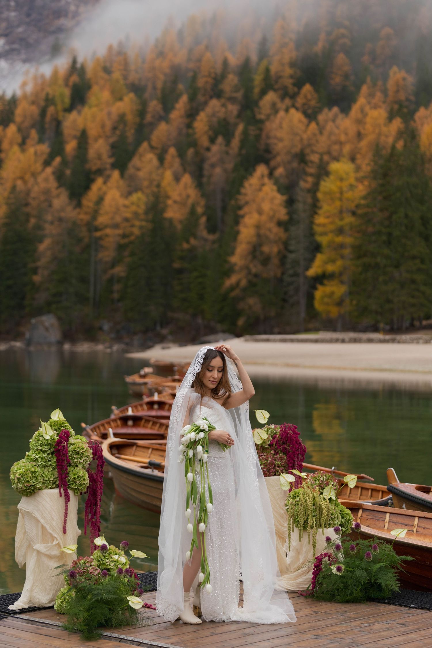 A bride stands on a dock with wooden boats and autumn trees reflecting in a serene mountain lake in the Dolomites