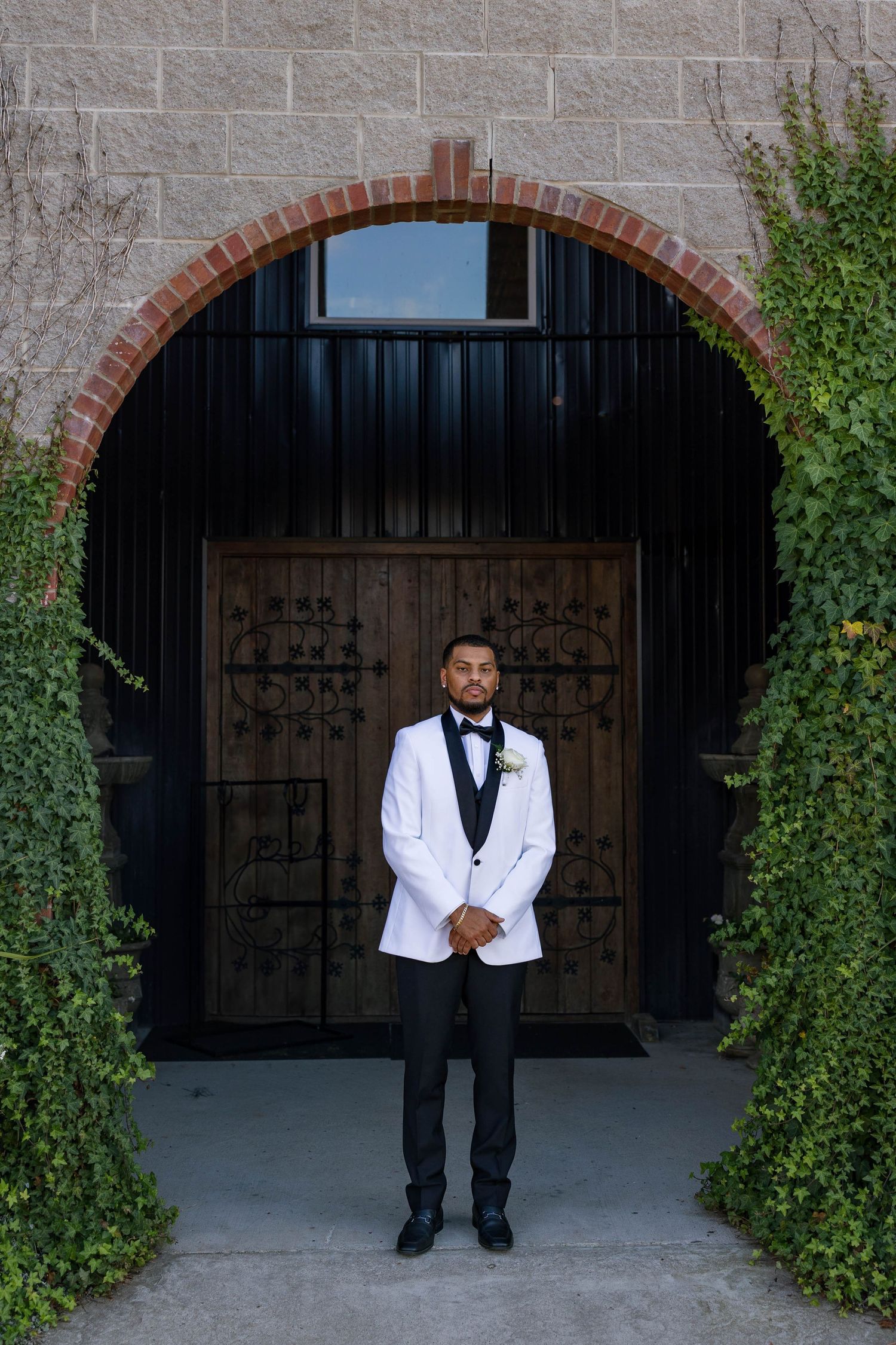 A groom in formal attire poses playfully in front of an arched stone doorway with ivy.