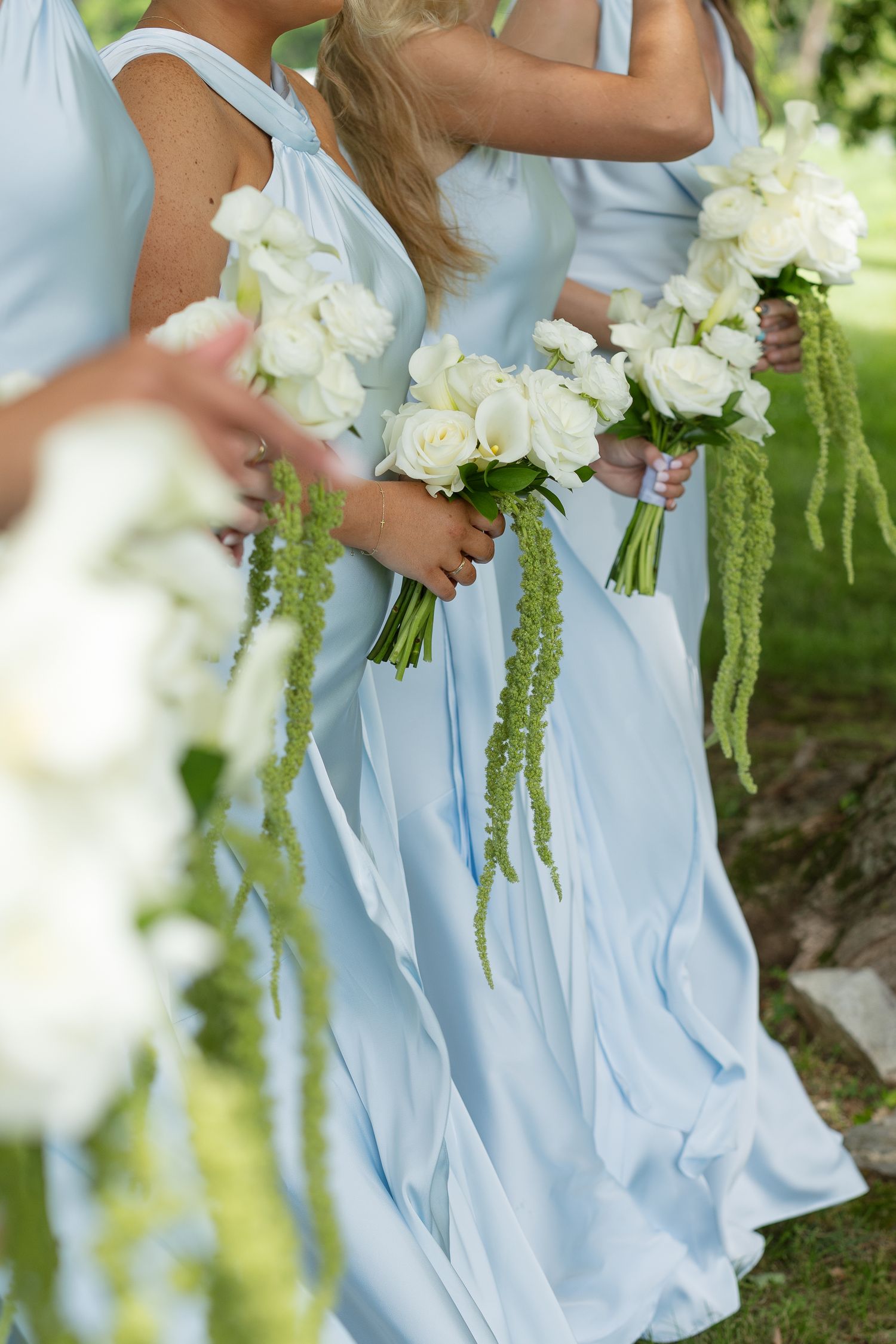 Bridesmaids in light blue gowns holding white rose bouquets with trailing green amaranthus at an outdoor wedding.