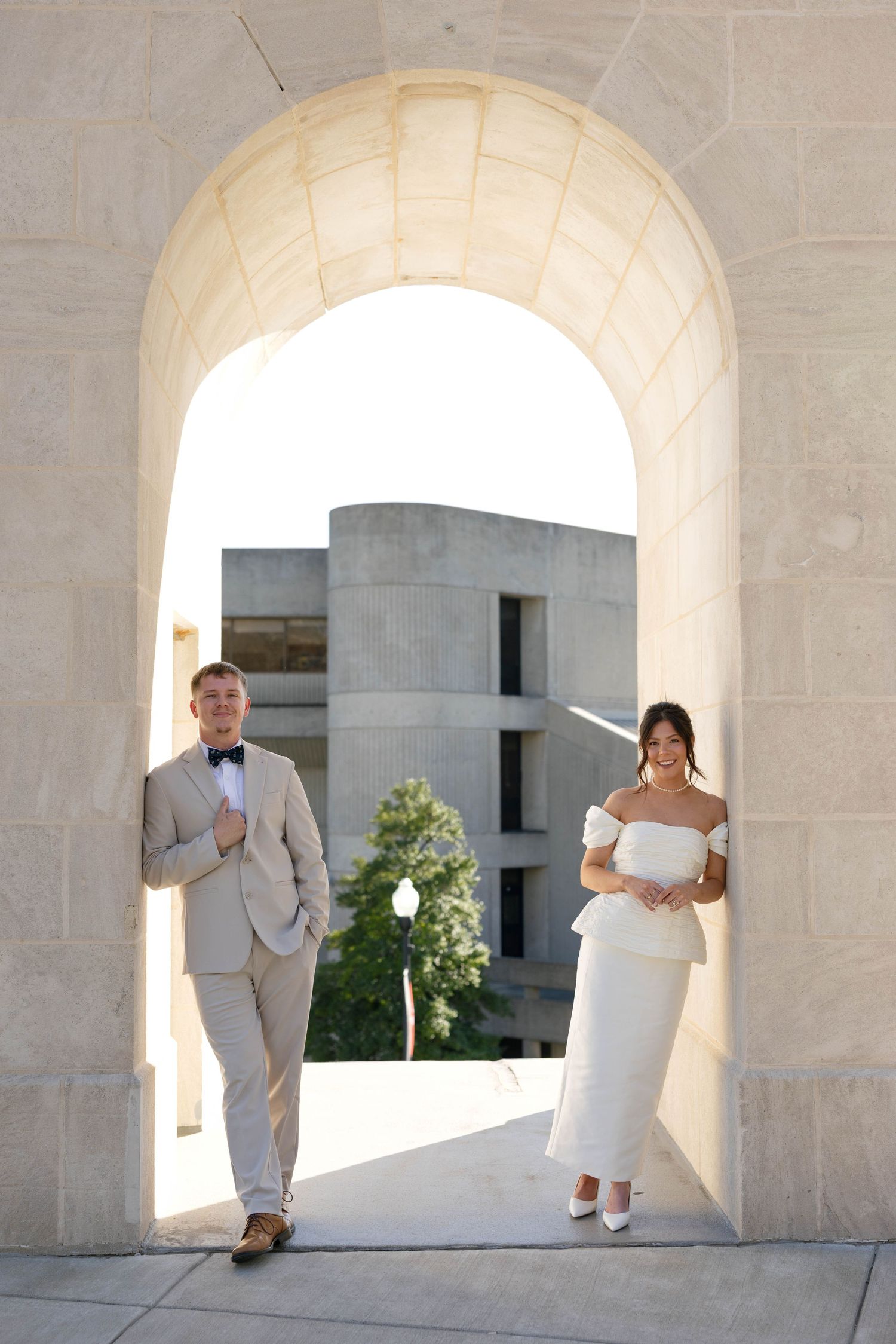 A couple in wedding attire poses under a stone arch, groom in beige suit, bride in white off-shoulder dress.