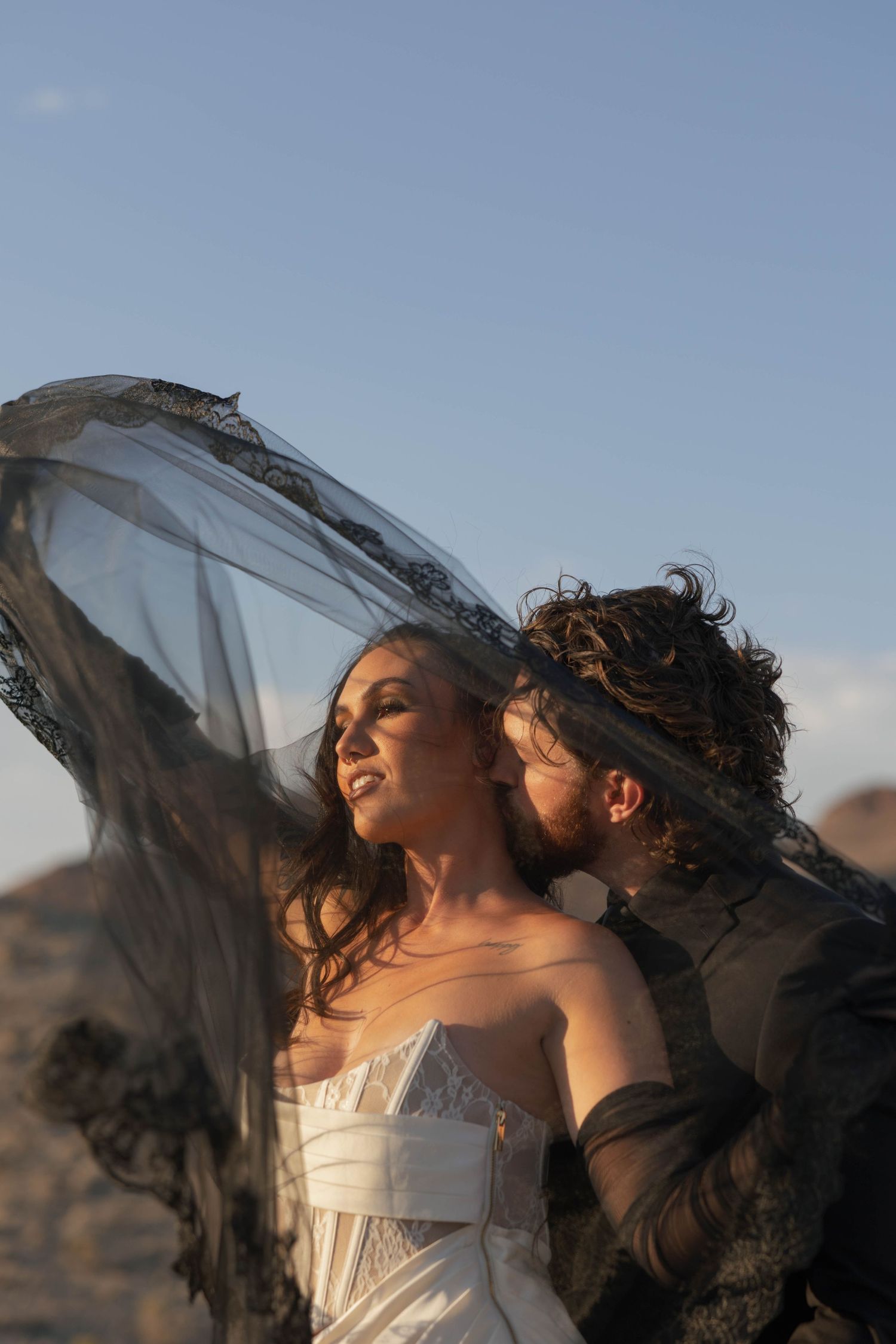 Bride in white gown with black lace veil blowing in wind, groom kissing her cheek in desert landscape.