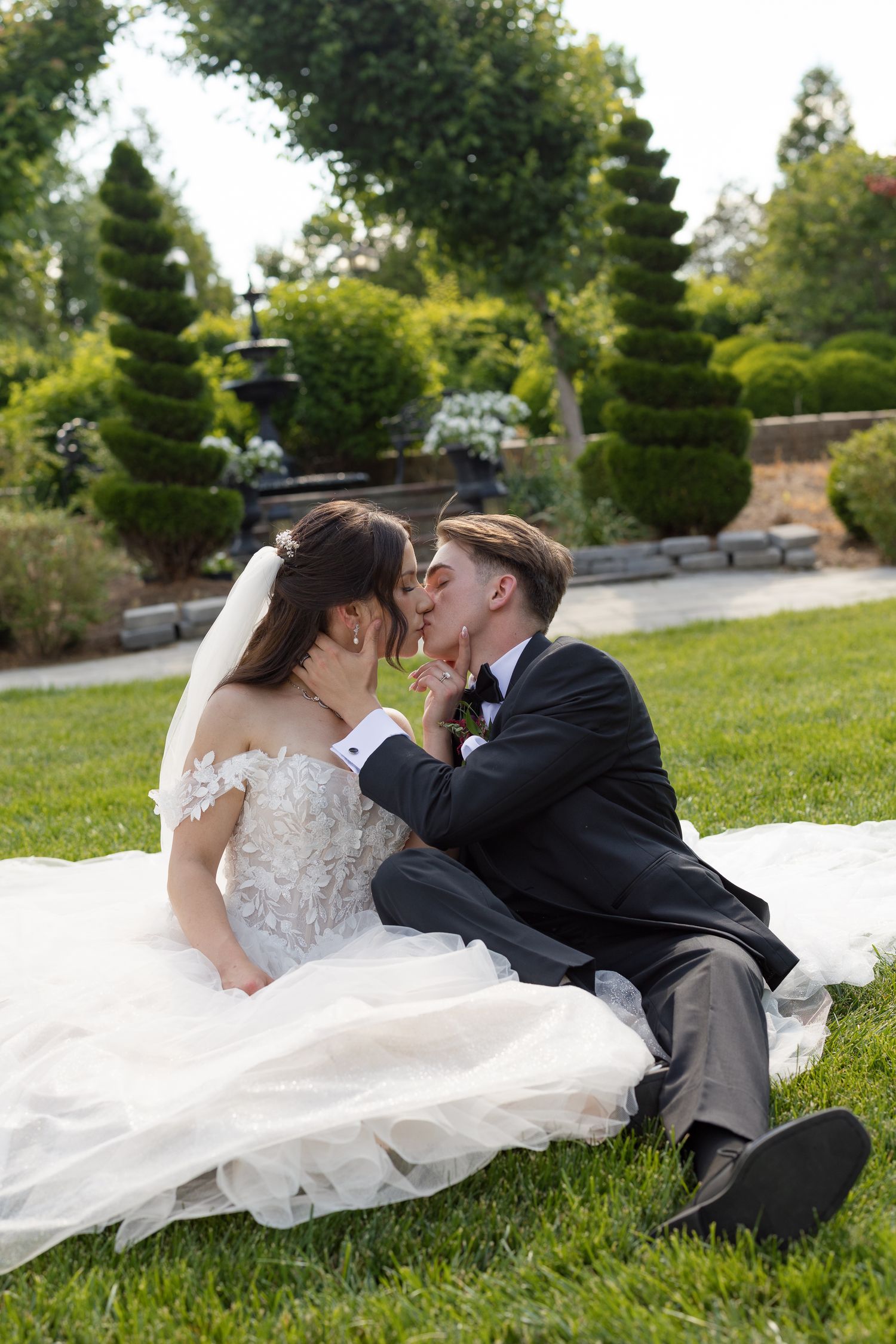 Bride and groom share a romantic kiss while sitting on green grass in a garden setting on their wedding day.