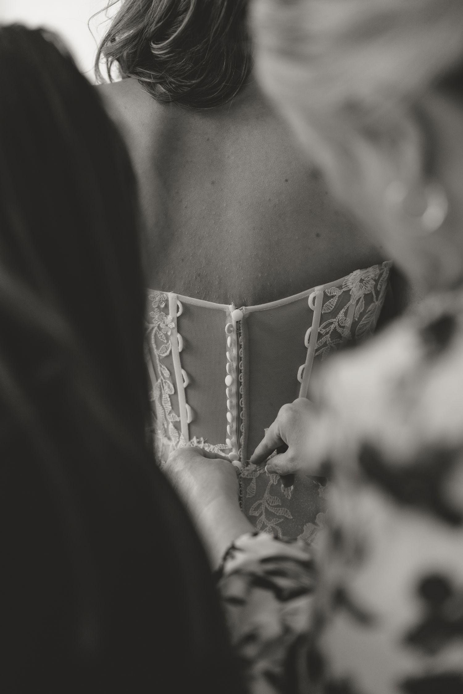 Black and white photo of a bride having her lace corset wedding dress buttoned up from behind by a bridesmaid.