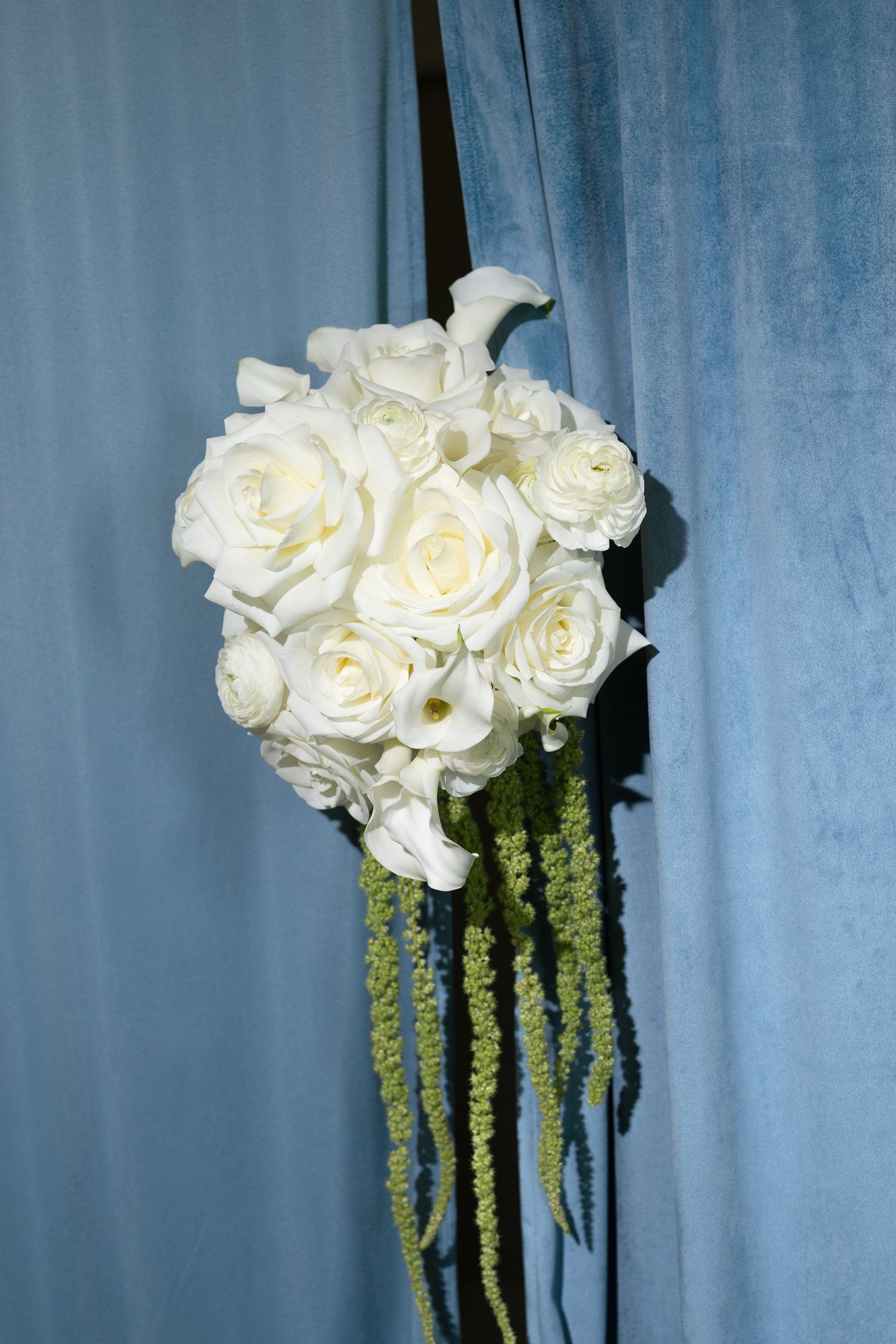 Elegant white rose and calla lily wedding bouquet with green amaranthus trails against a blue curtain backdrop.