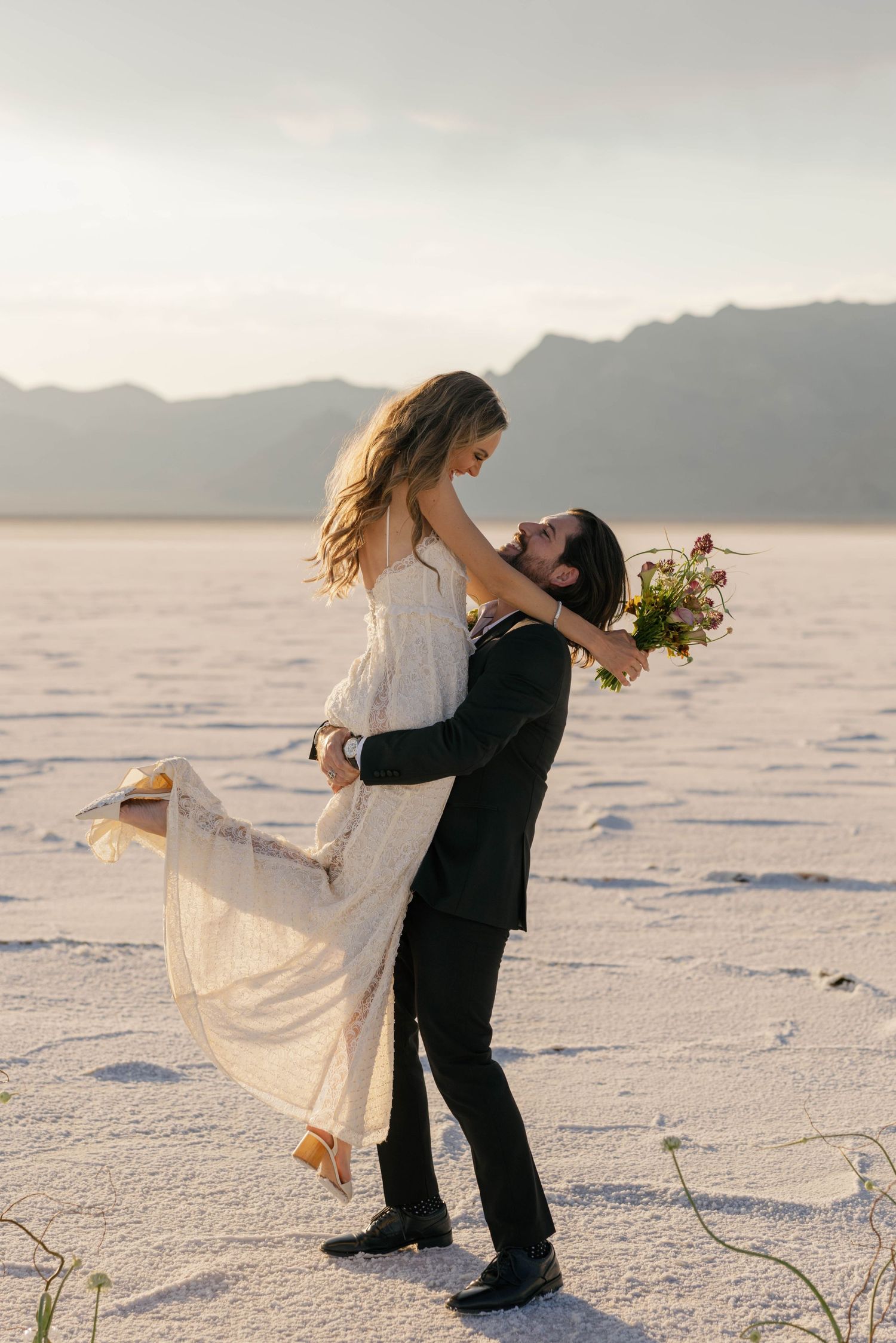 A couple in wedding attire dances together on white salt flats under cloudy skies.