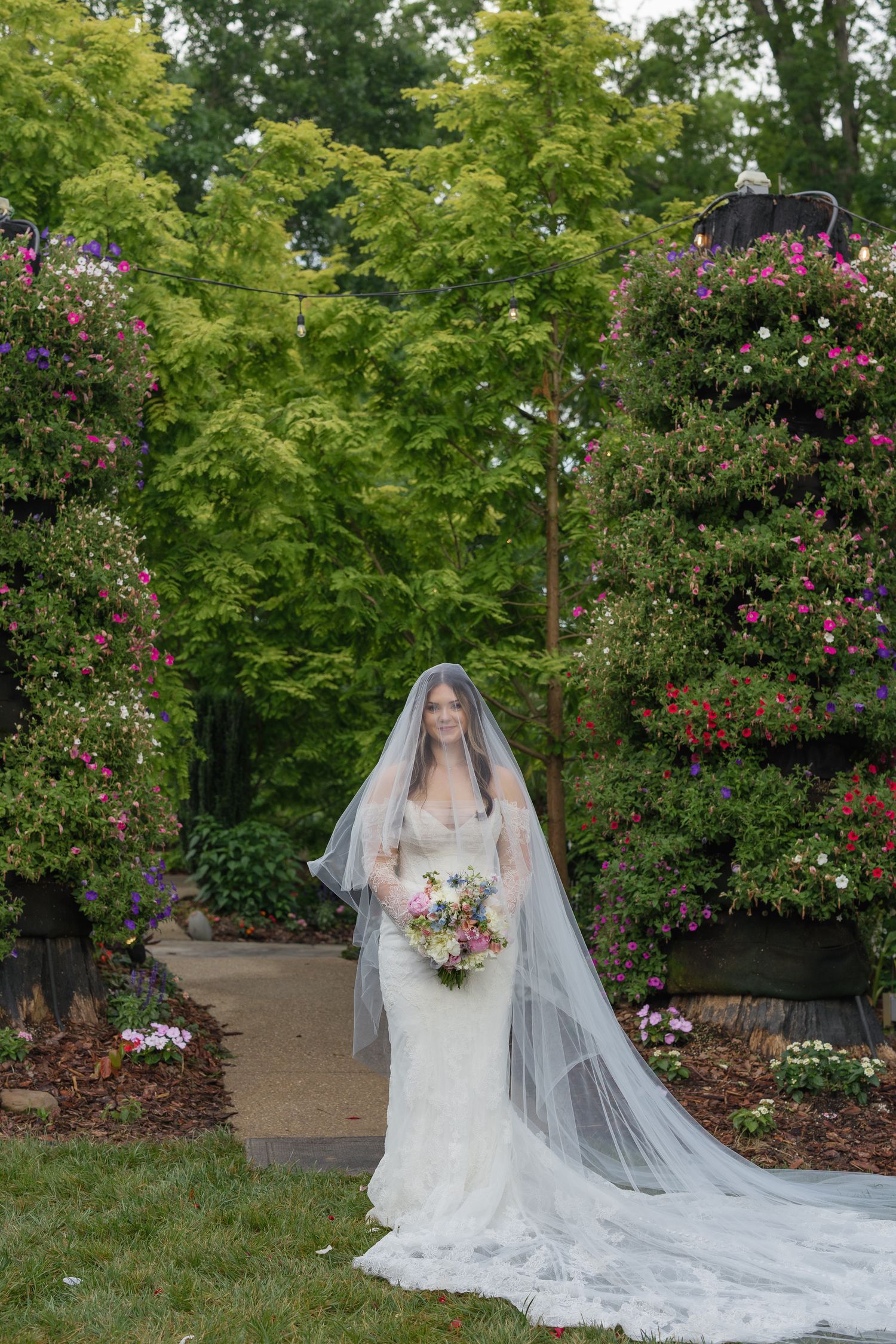 Bride in lace gown and long veil holds colorful bouquet, standing in a lush garden with blooming flowers.