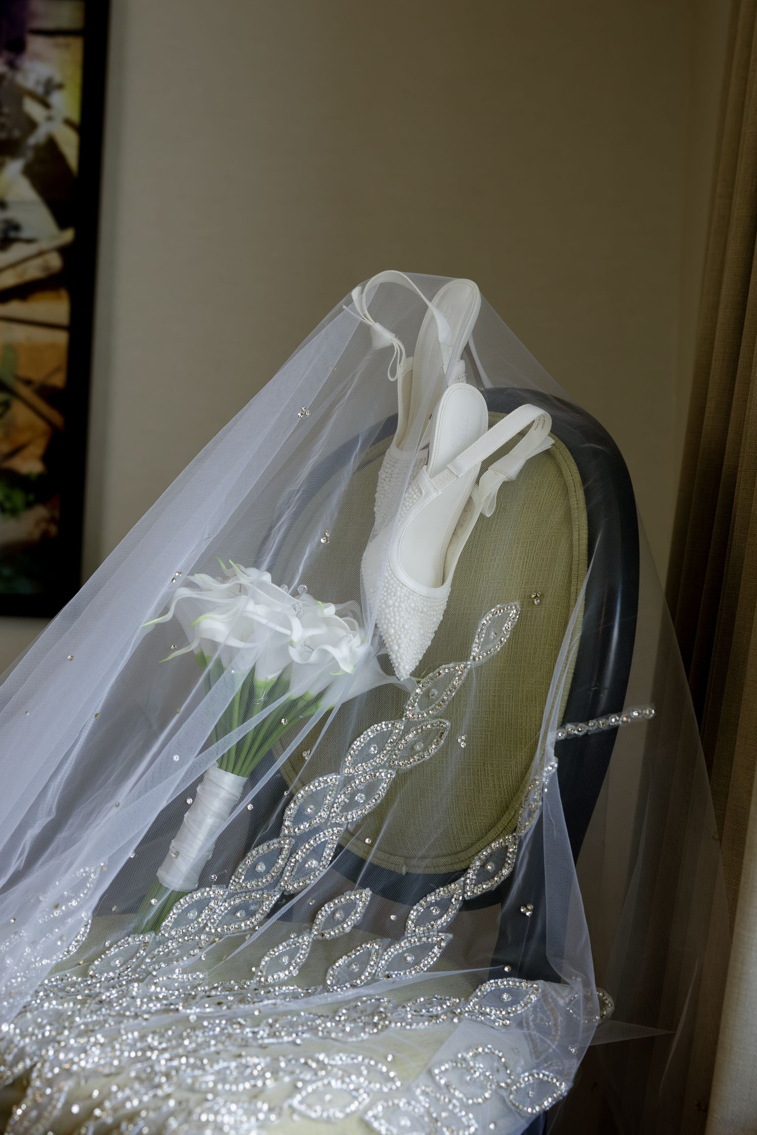 Elegant bridal veil with beaded lace trim draped over a chair beside white calla lily bouquet and wedding shoes.