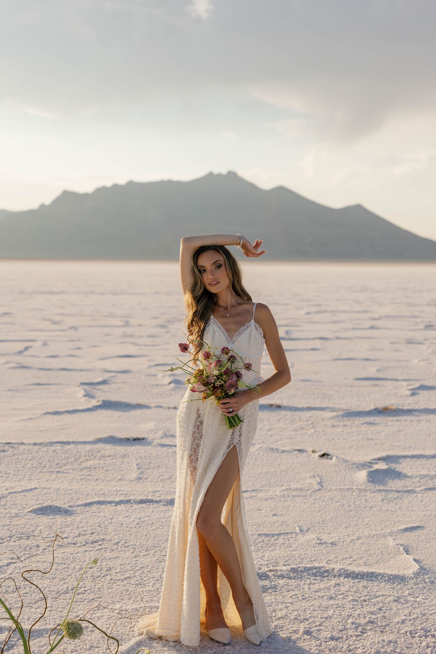A bride in a flowing white dress poses elegantly on salt flats at sunset with mountains in the background.