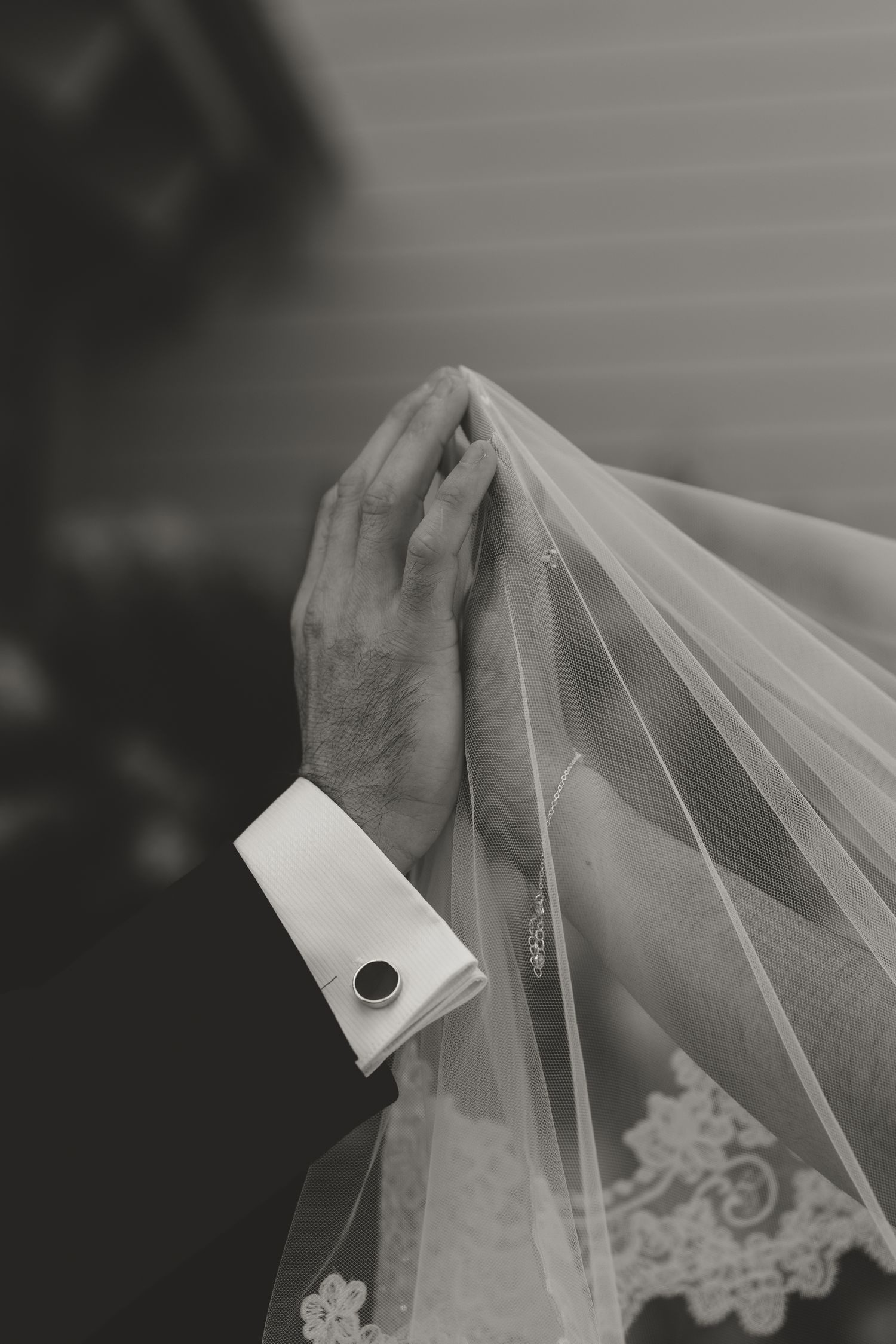 Black and white photo of a groom's hand holding a bride's veil, wearing a cufflink, at a wedding ceremony.