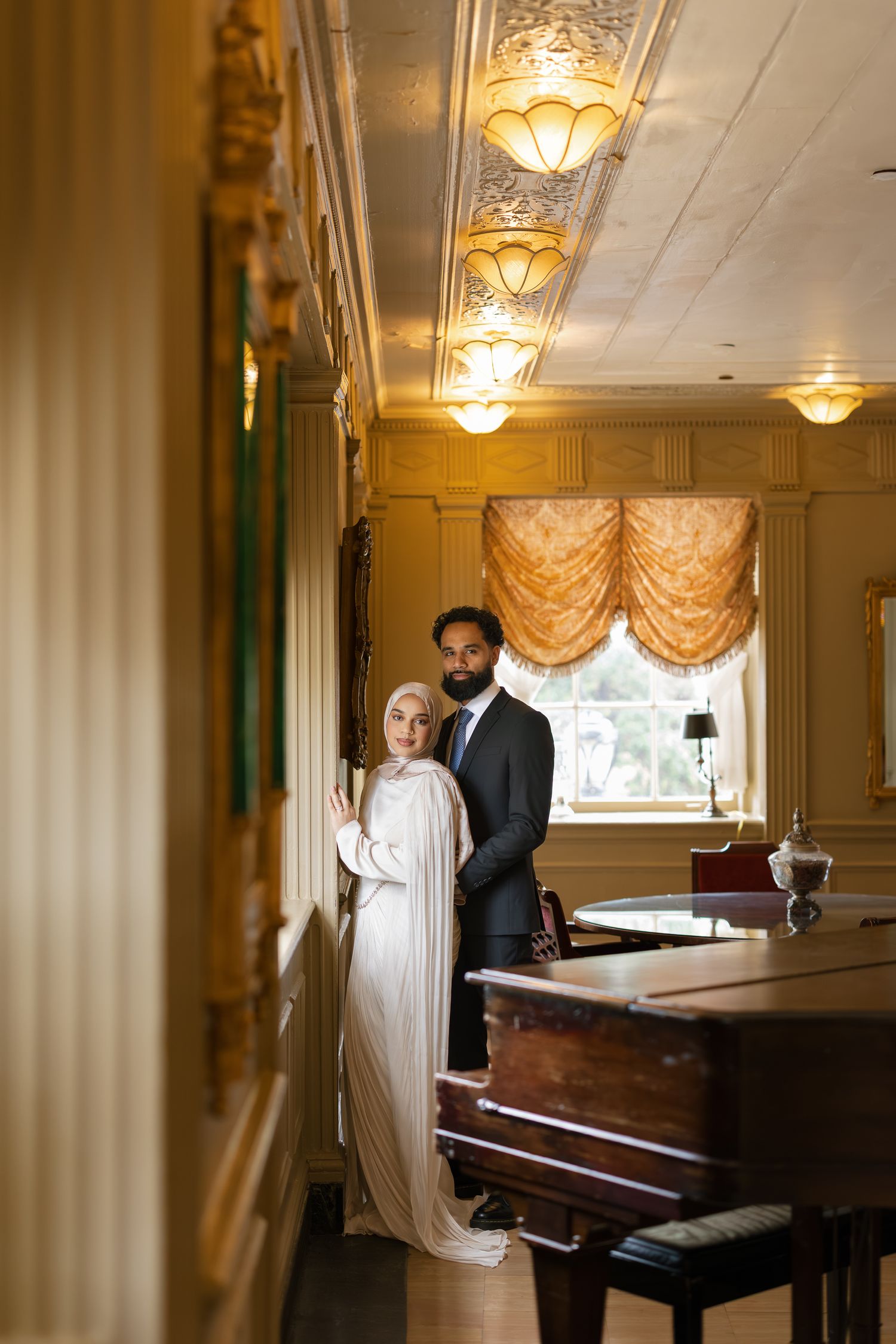 Elegant couple in formal attire poses near a grand piano in a luxurious golden room with ornate ceiling lights.