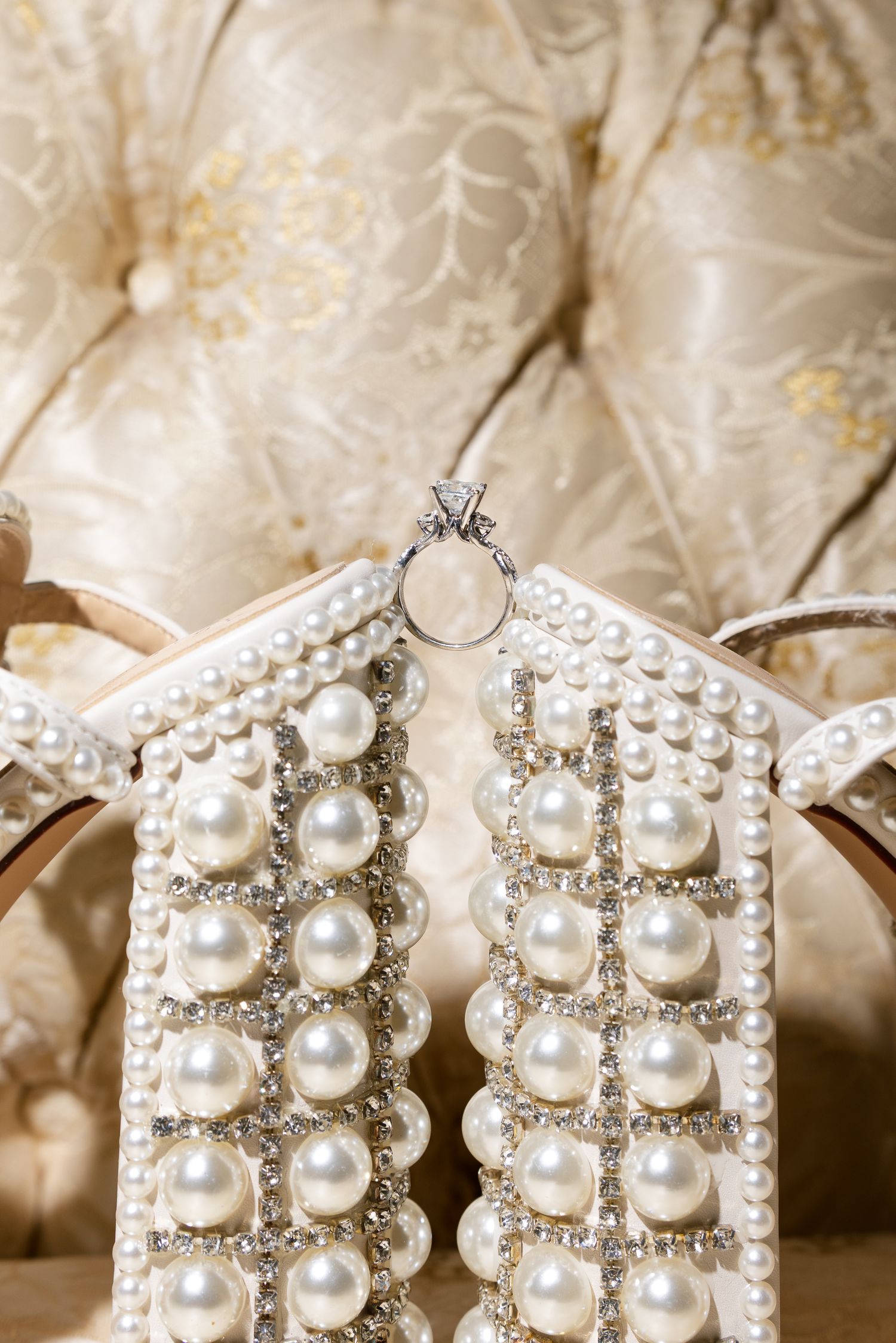 Pearl and crystal embellished bridal heels displayed with a diamond engagement ring on ivory tufted fabric backdrop.