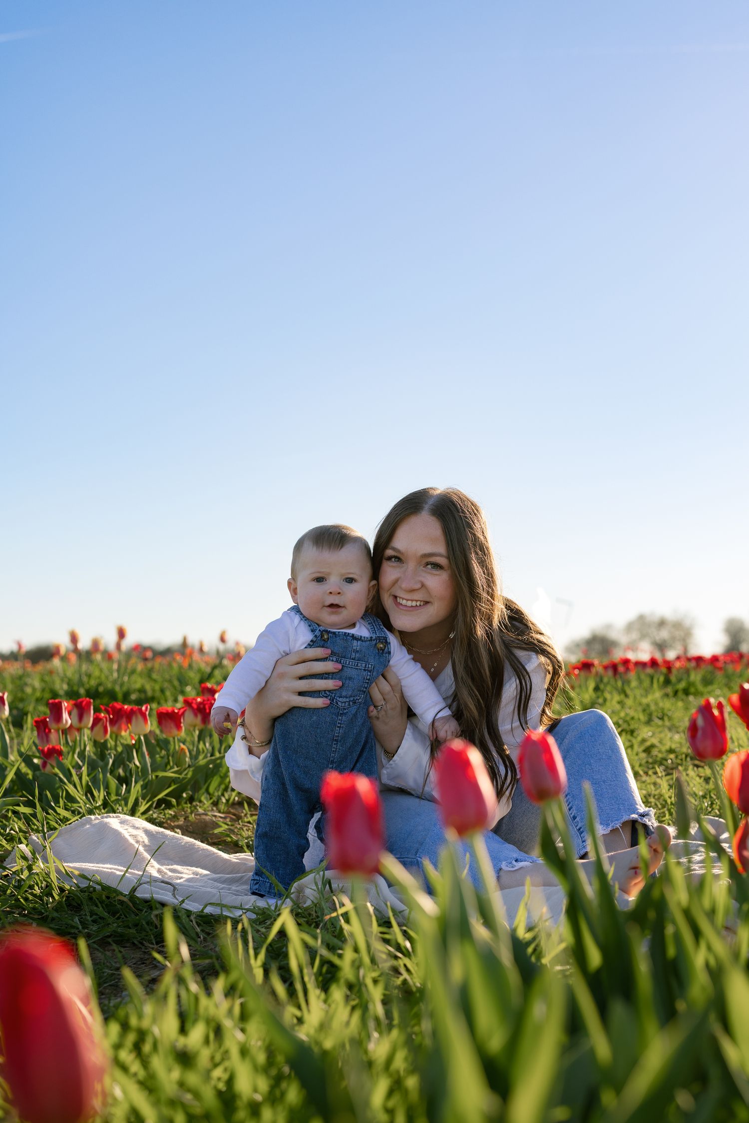 A smiling young woman holds a baby in a vibrant red tulip field under a clear blue sky.