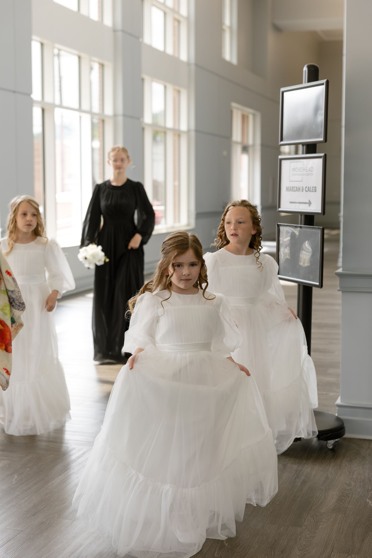 Three young girls in white flower girl dresses walk through a wedding venue hall, with a woman in black standing behind them.