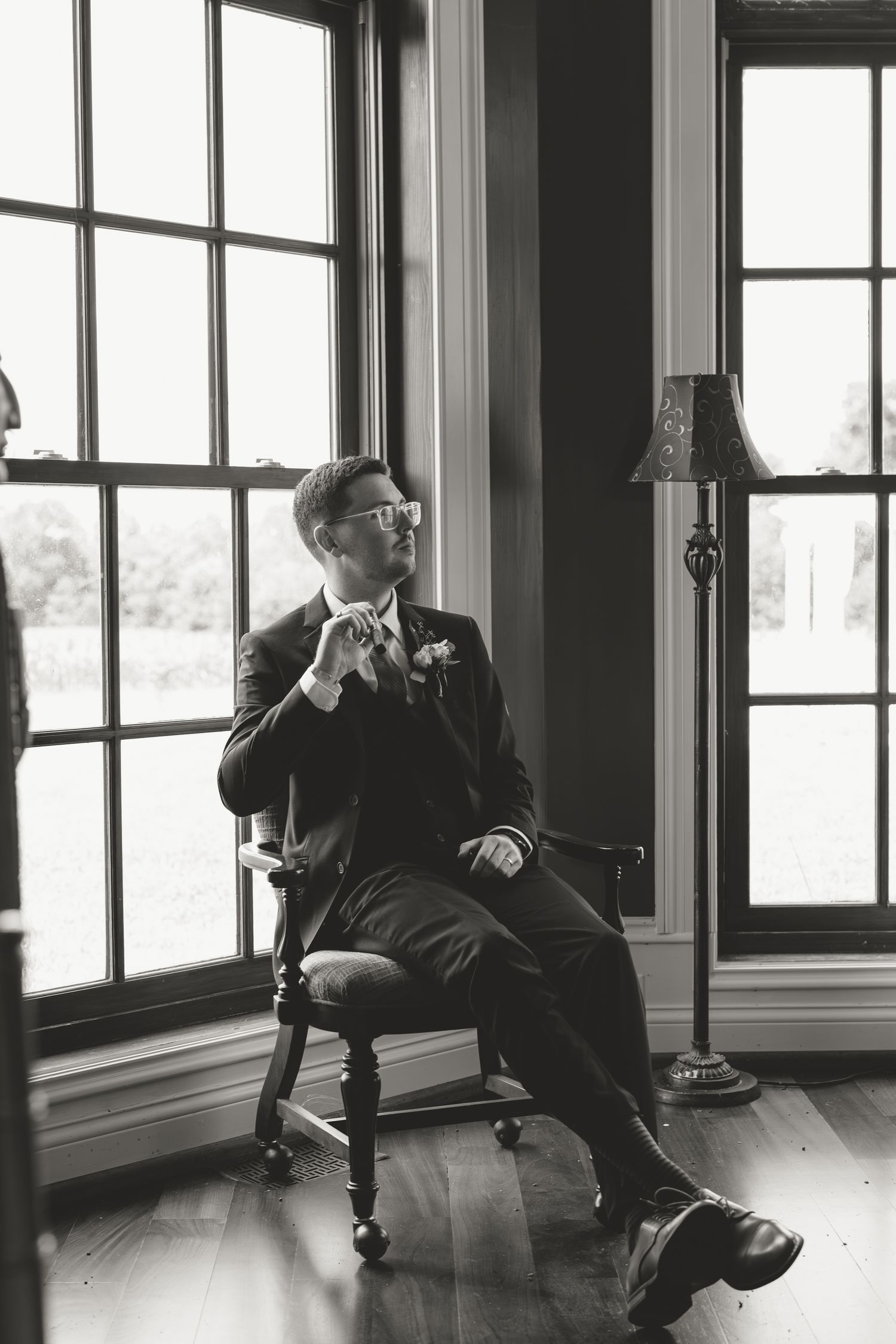 Black and white photo of a groom in a suit adjusting his tie while seated in a wooden chair by large windows.