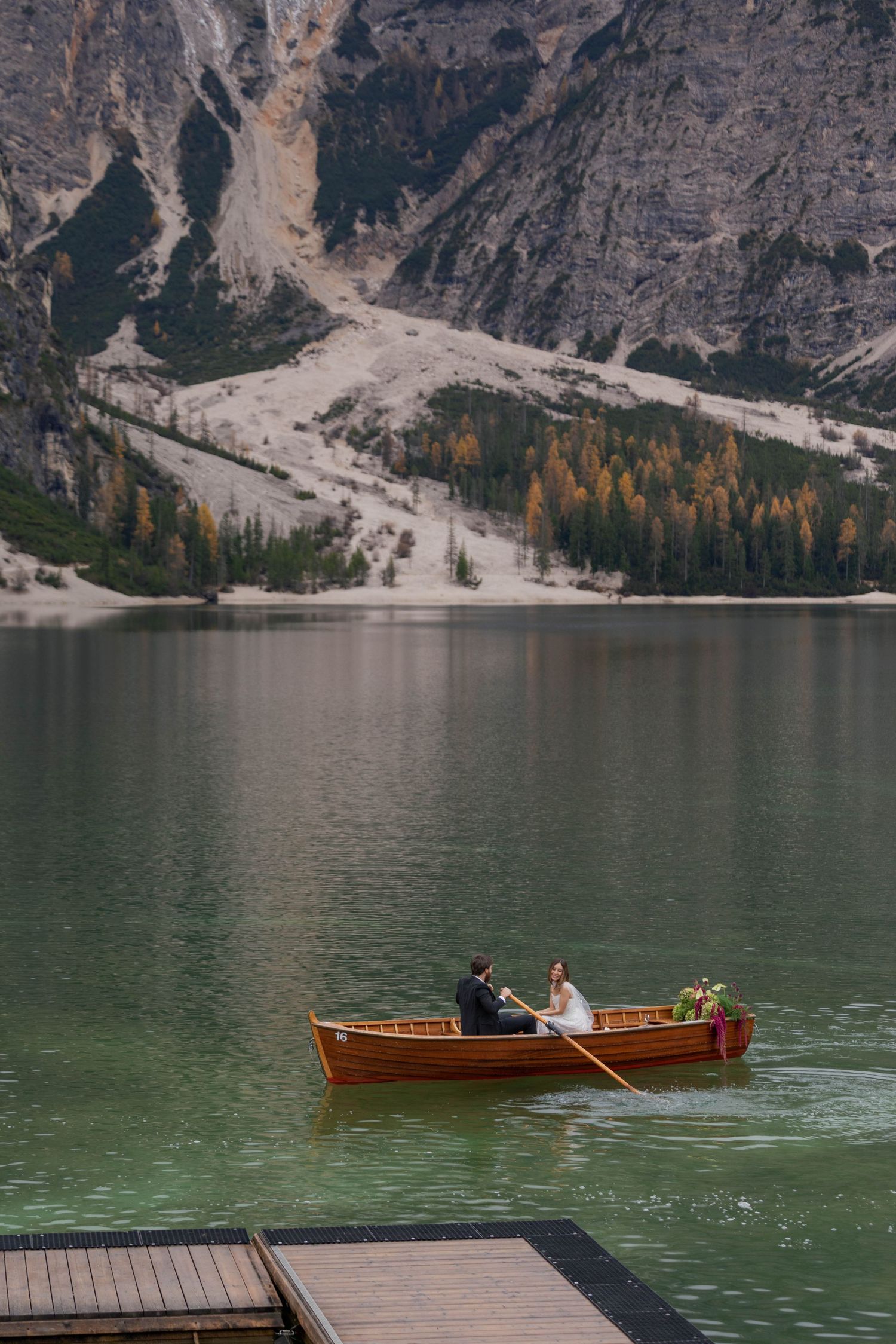Couple in wedding attire rowing a wooden boat on a serene alpine lake with mountain backdrop.