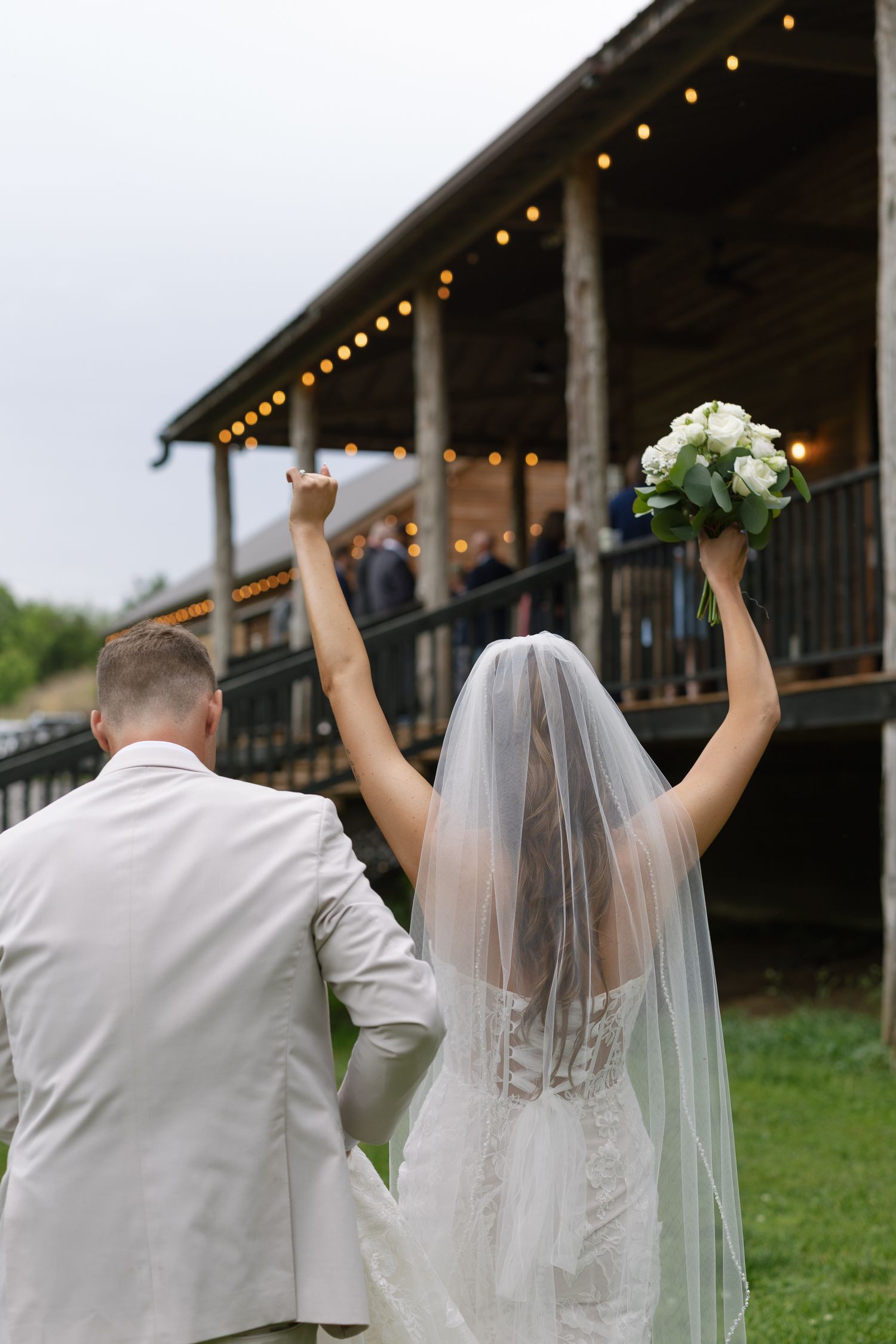 Bride and groom raising arms in celebration walking toward a rustic venue with string lights on the porch.