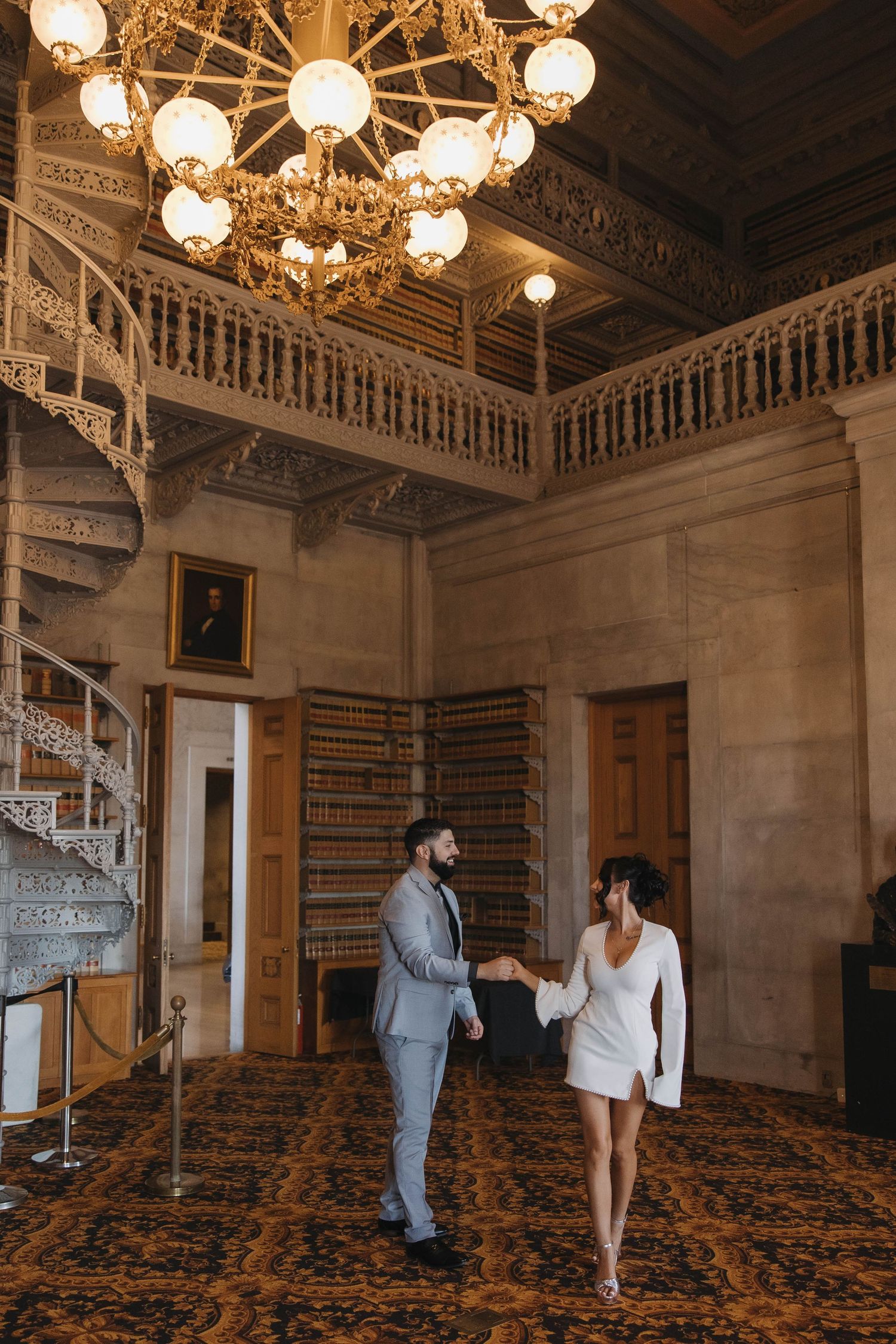 Couple in wedding attire holding hands in an ornate library with grand chandelier and spiral staircase.