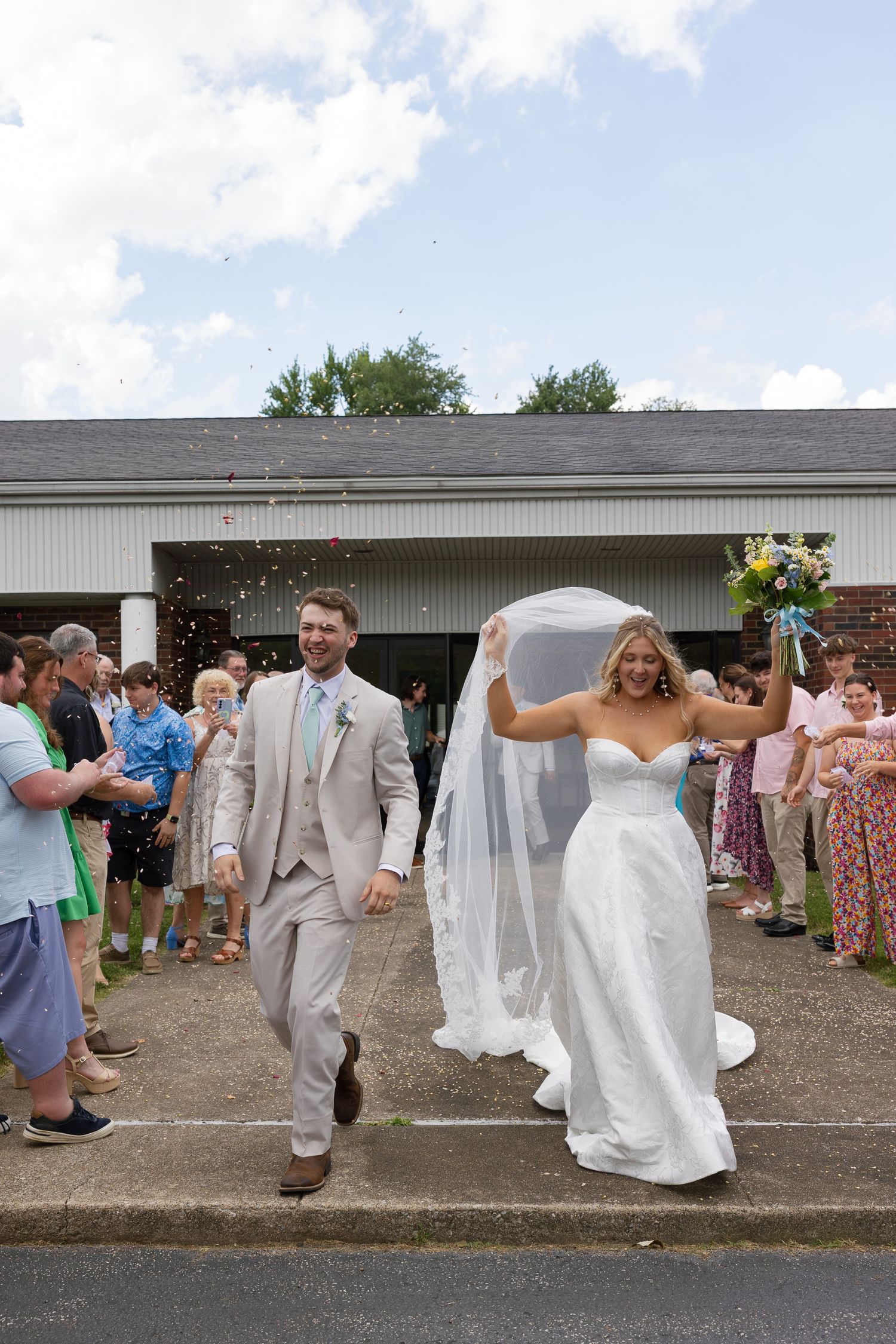 Joyful newlyweds exit wedding ceremony as guests celebrate with confetti, bride in white gown holding bouquet, groom in tan suit.