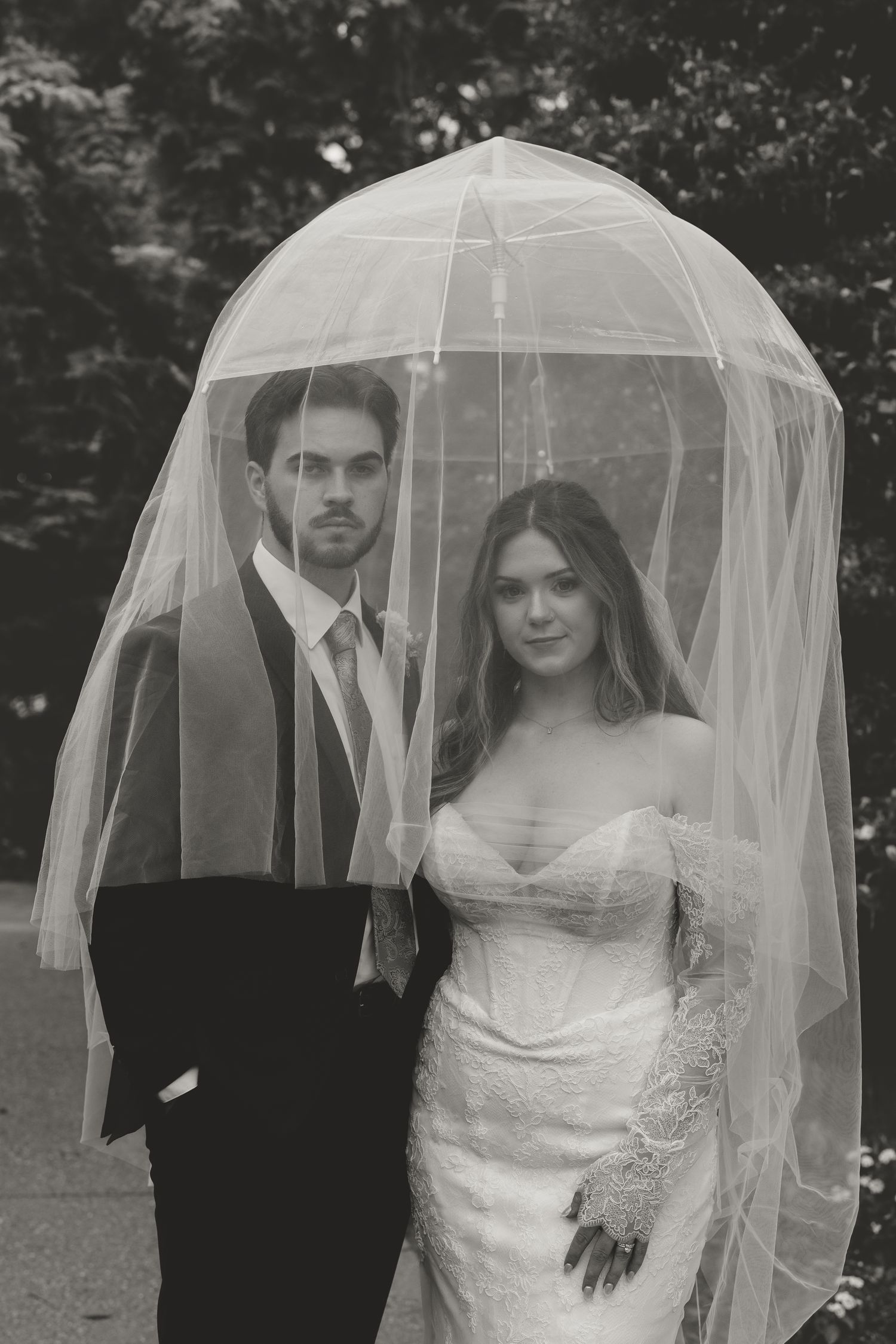 Black and white wedding photo of bride and groom posing together under a flowing veil outdoors.