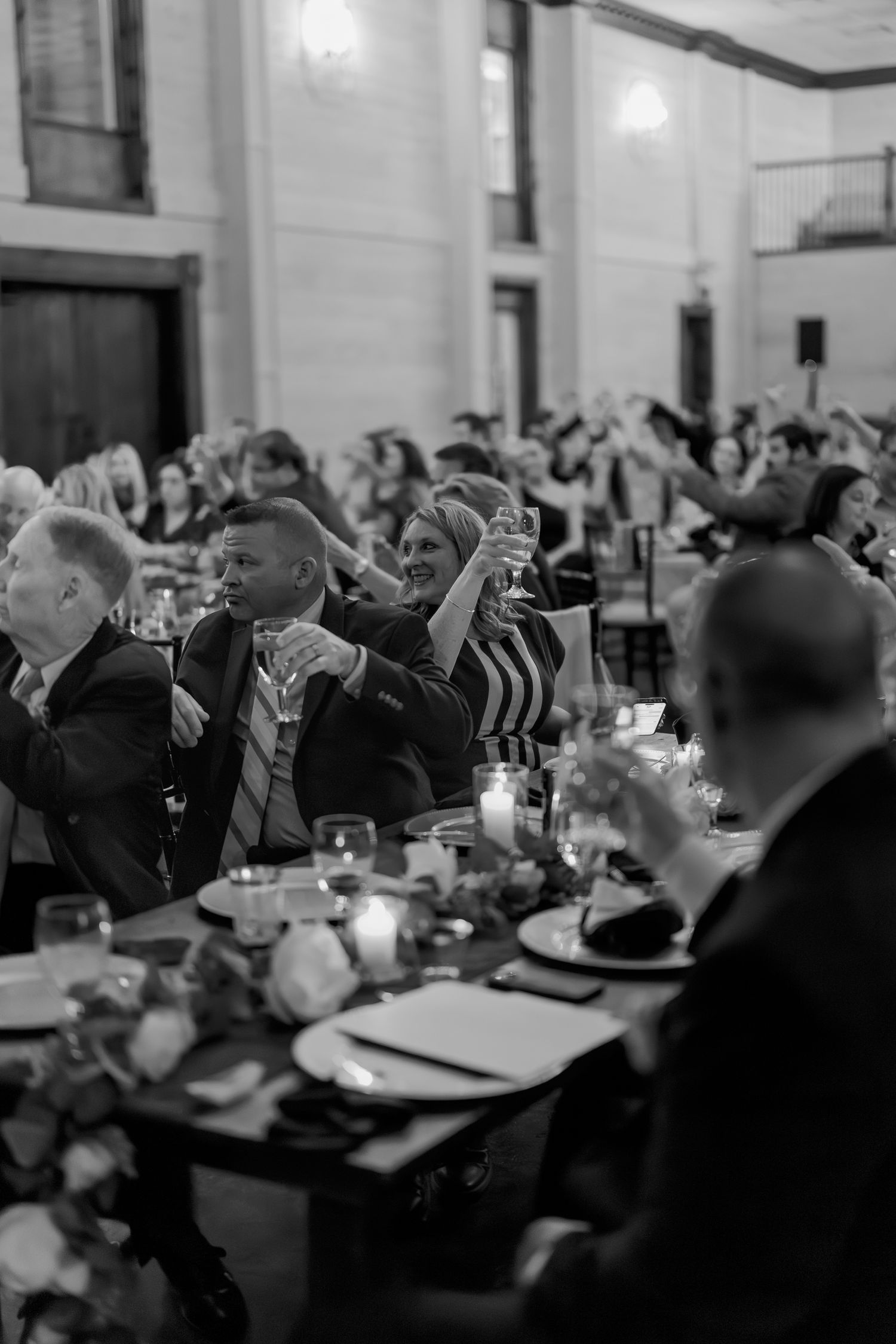 Black and white photo of wedding reception guests raising glasses for a toast in an elegant ballroom.