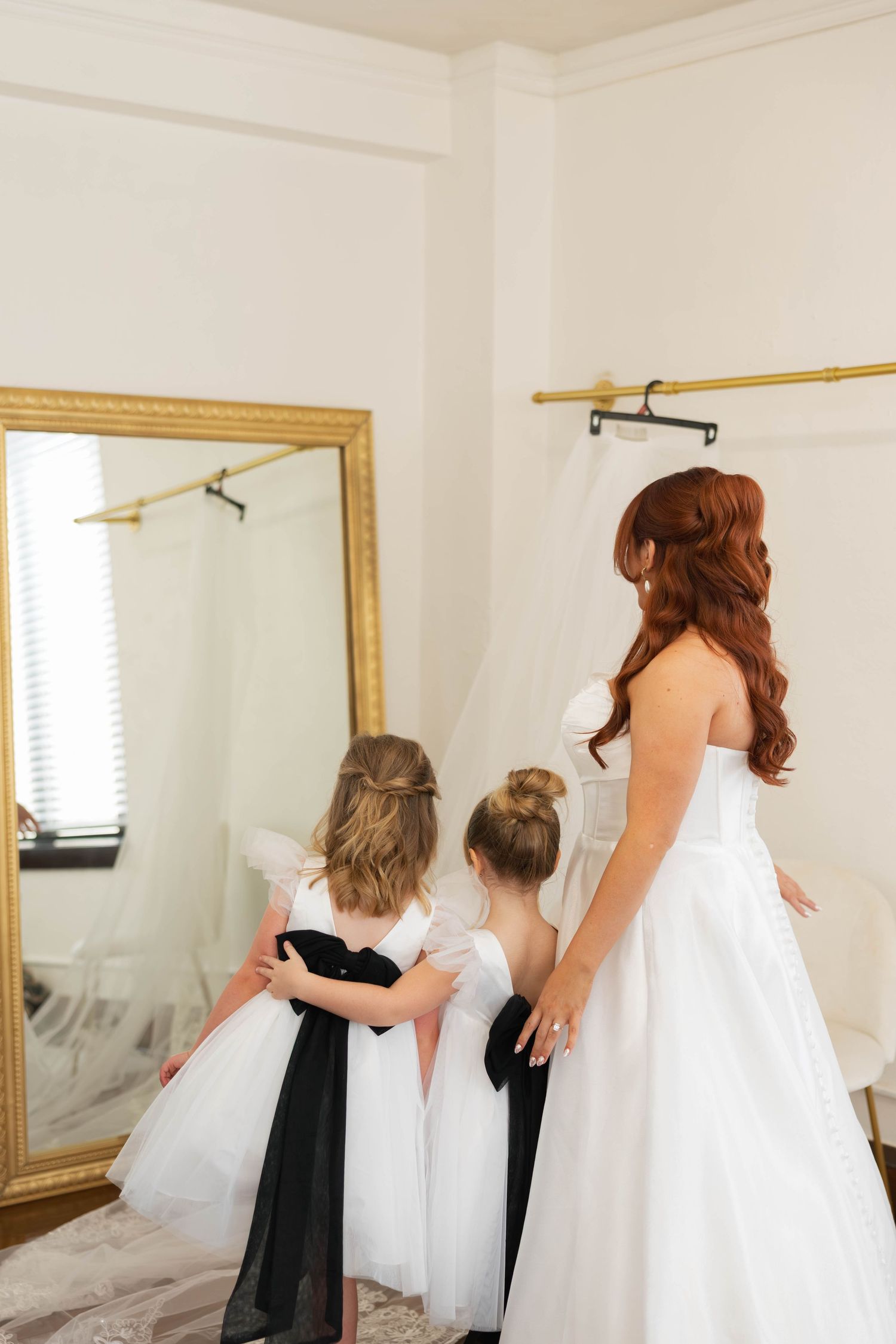 Wedding dress fitting with flower girls in matching white and black dresses looking in a mirror.