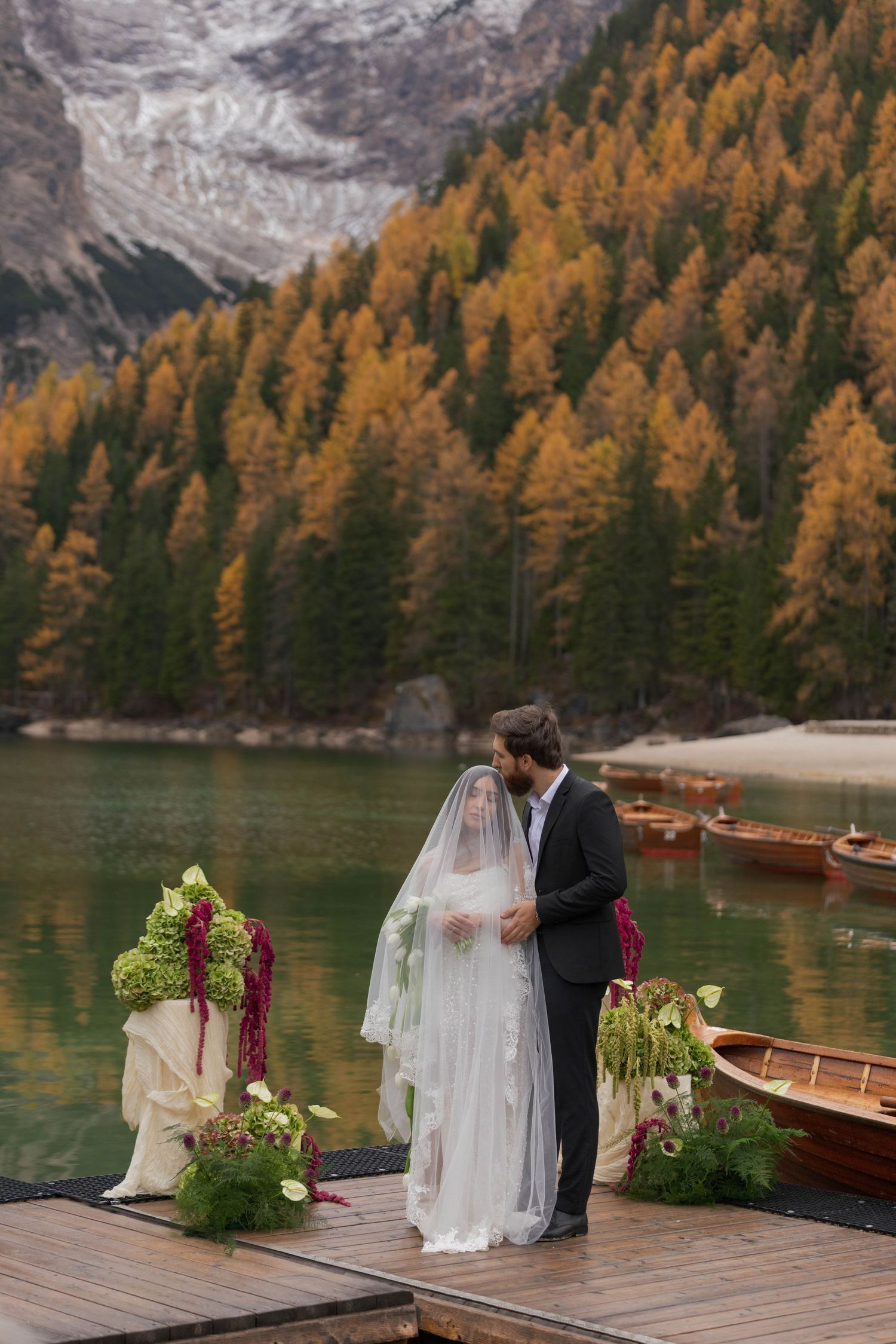 Couple kissing at outdoor lakeside wedding ceremony on dock with autumn foliage and mountain backdrop.