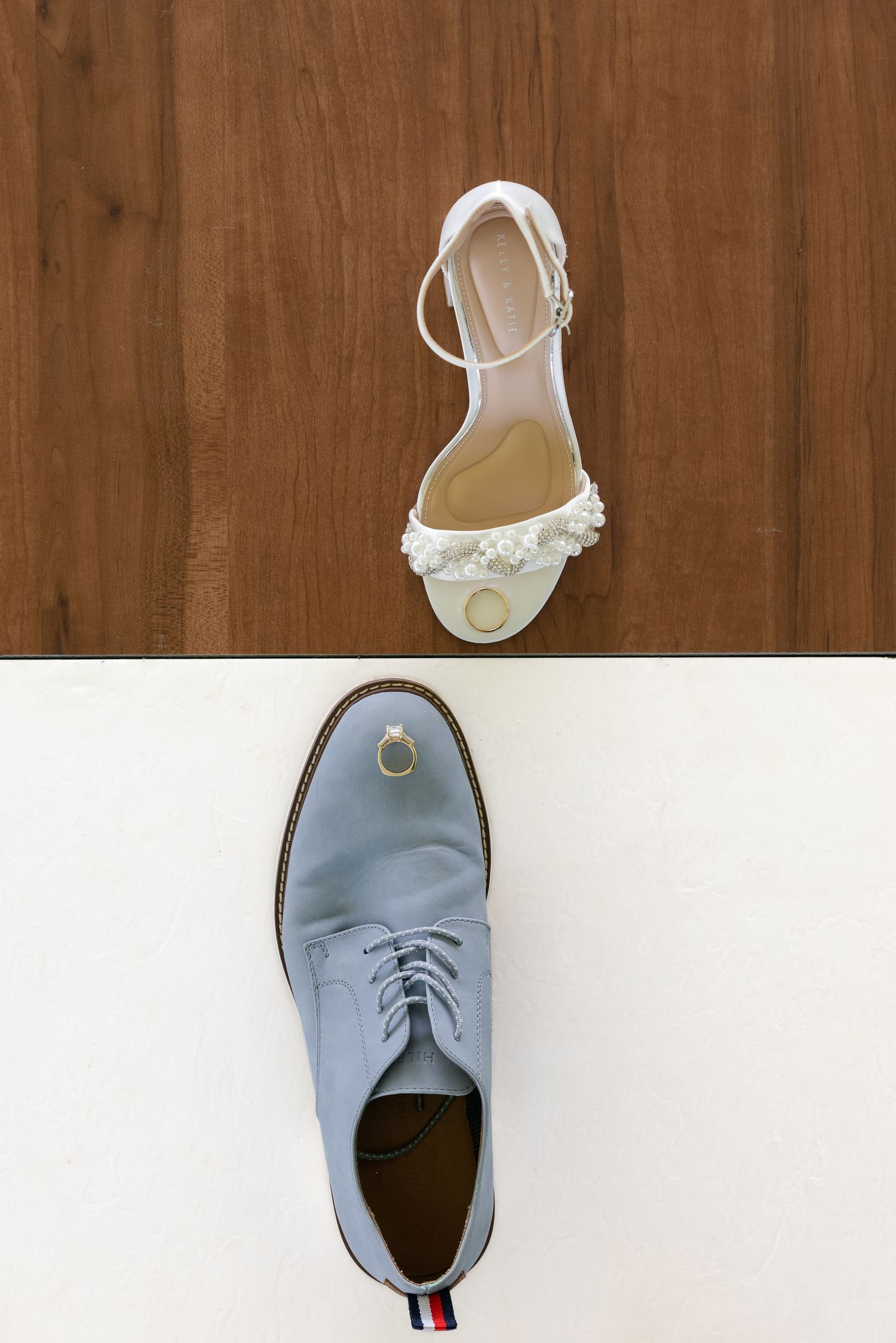 White bridal sandal with floral detail and gray groom dress shoe displayed with gold wedding rings on wood and white surfaces.