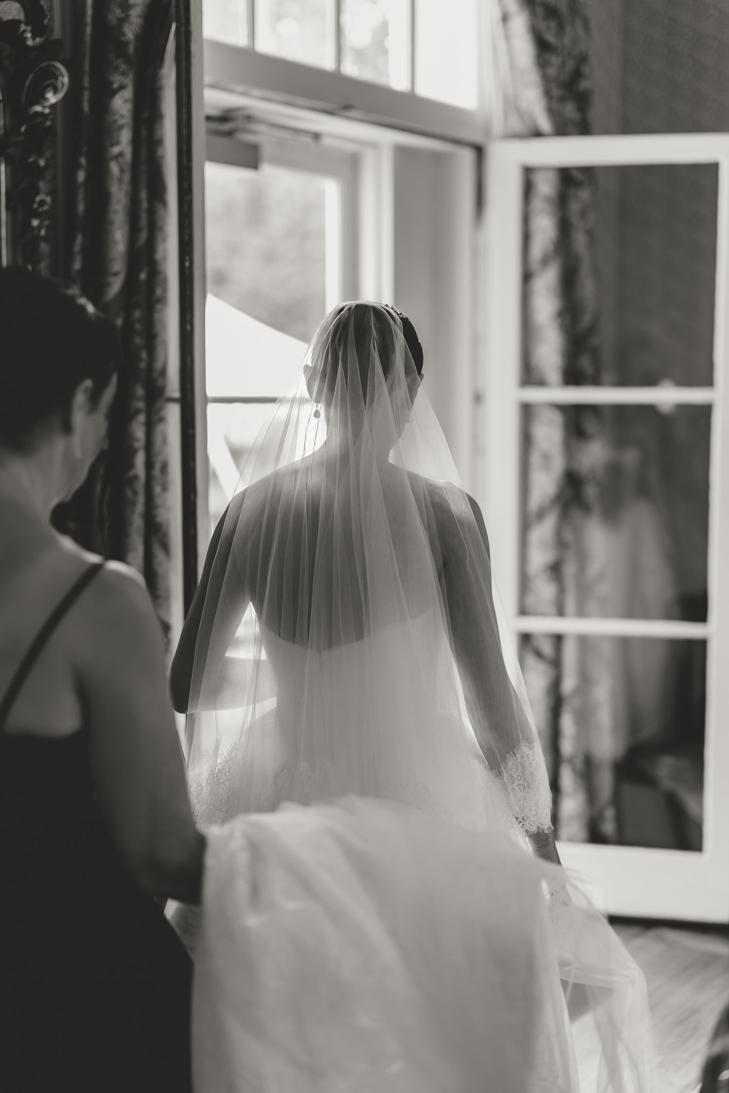Black and white photo of a bride in a wedding gown and veil standing by a window with a bridesmaid beside her.