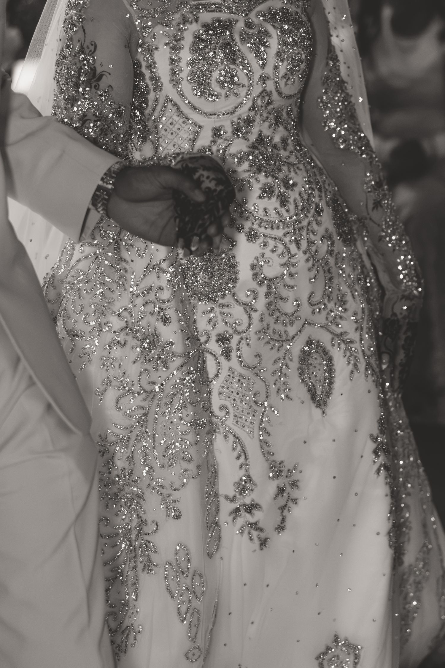 Black and white photo of a bride in a heavily beaded and embroidered wedding gown held by a groom in a white suit.