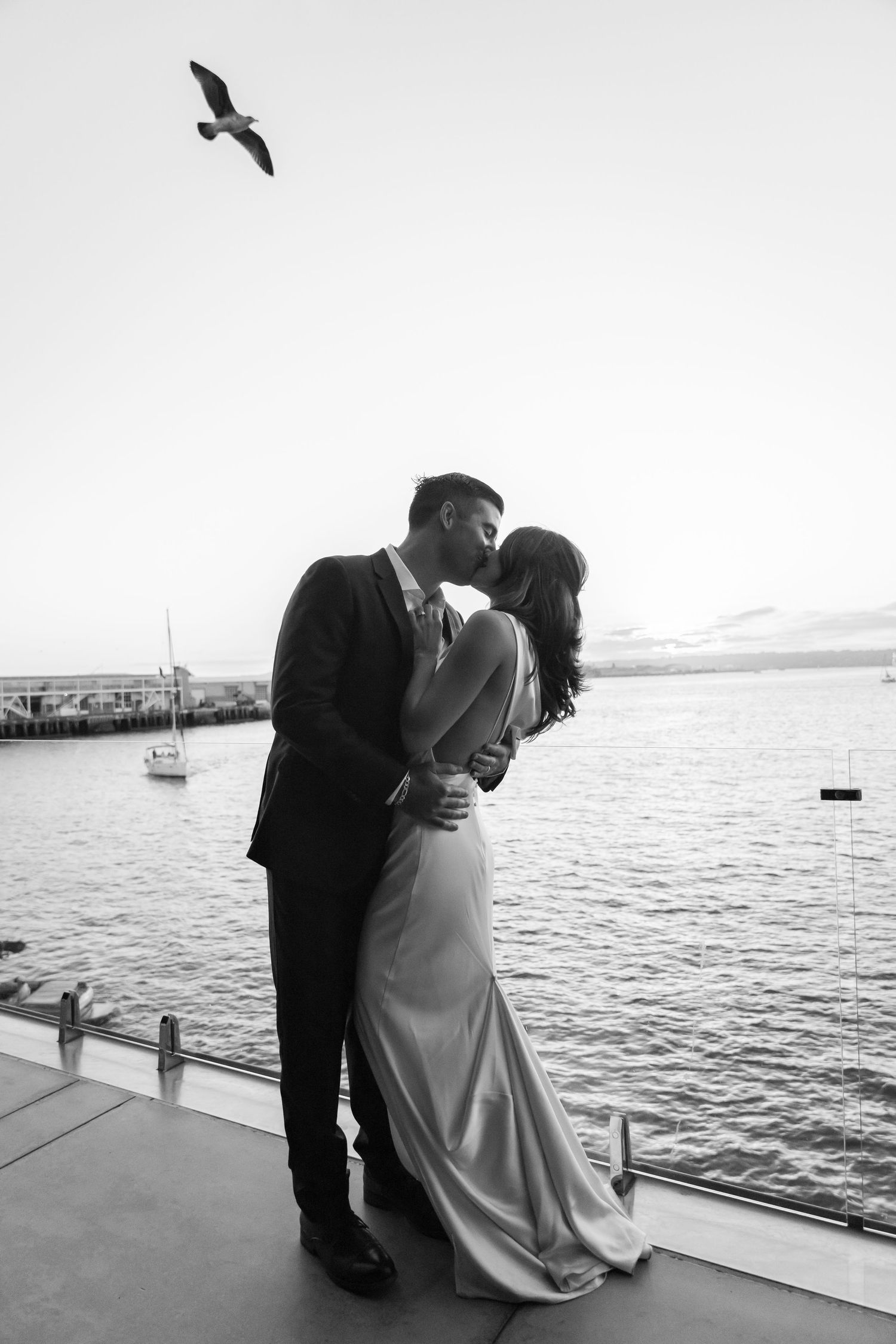 A romantic black and white wedding photo of a couple embracing on a dock by the water with a bird flying overhead.