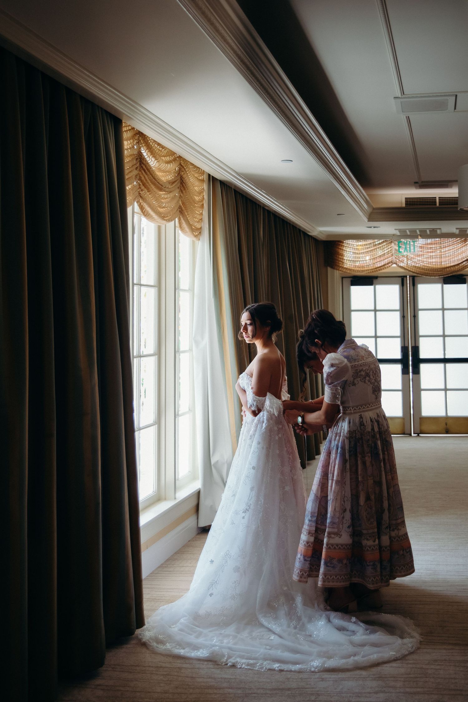Wedding scene shows a bride in flowing white gown being helped with final preparations in an elegant room.