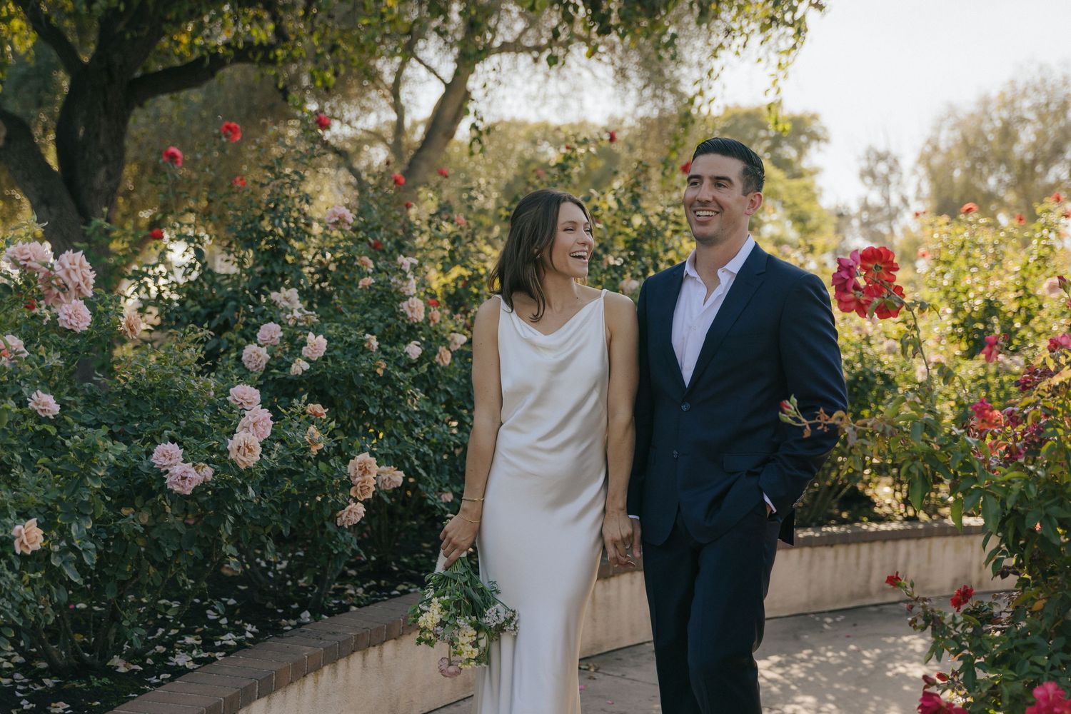 A couple shares a romantic moment in a blooming garden pathway with sunlight filtering through the trees.