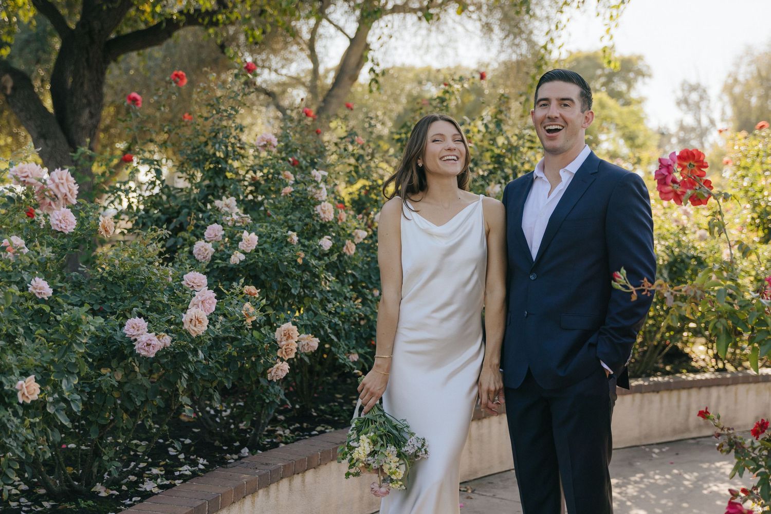 A couple stands together in a lush garden setting with blooming roses and greenery in the background.