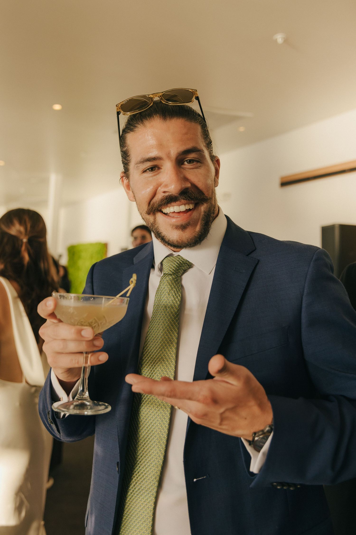 Man in navy suit and green tie holding up cocktail glasses at formal event.