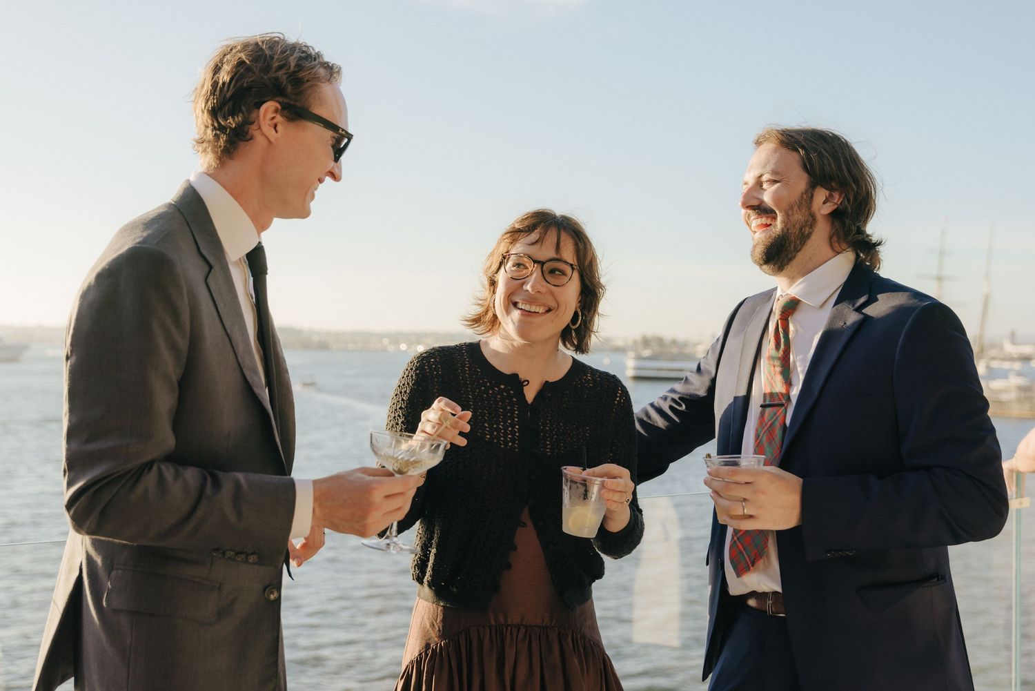 Wedding party laughs together while posing for casual photos by the waterfront on a sunny day.