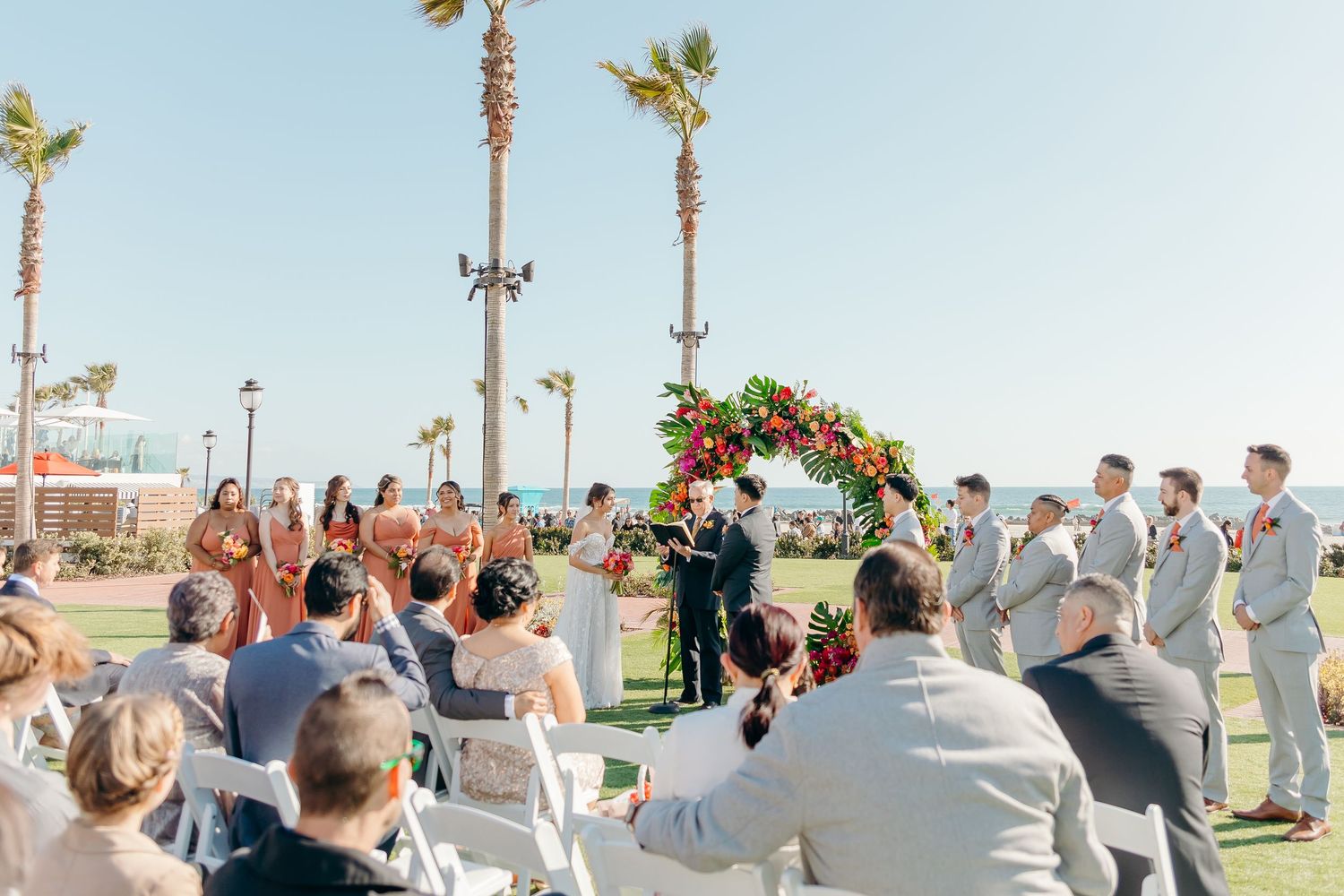 Outdoor beachside wedding ceremony with palm trees and guests seated in white chairs on a sunny day.