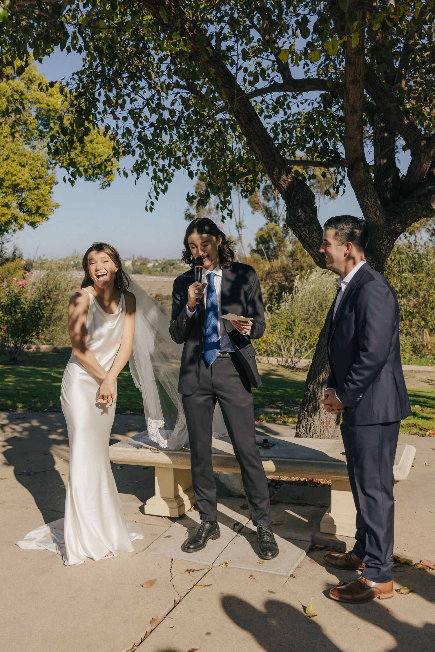 Wedding party shares a candid laughing moment together outdoors under a large tree on a sunny day.