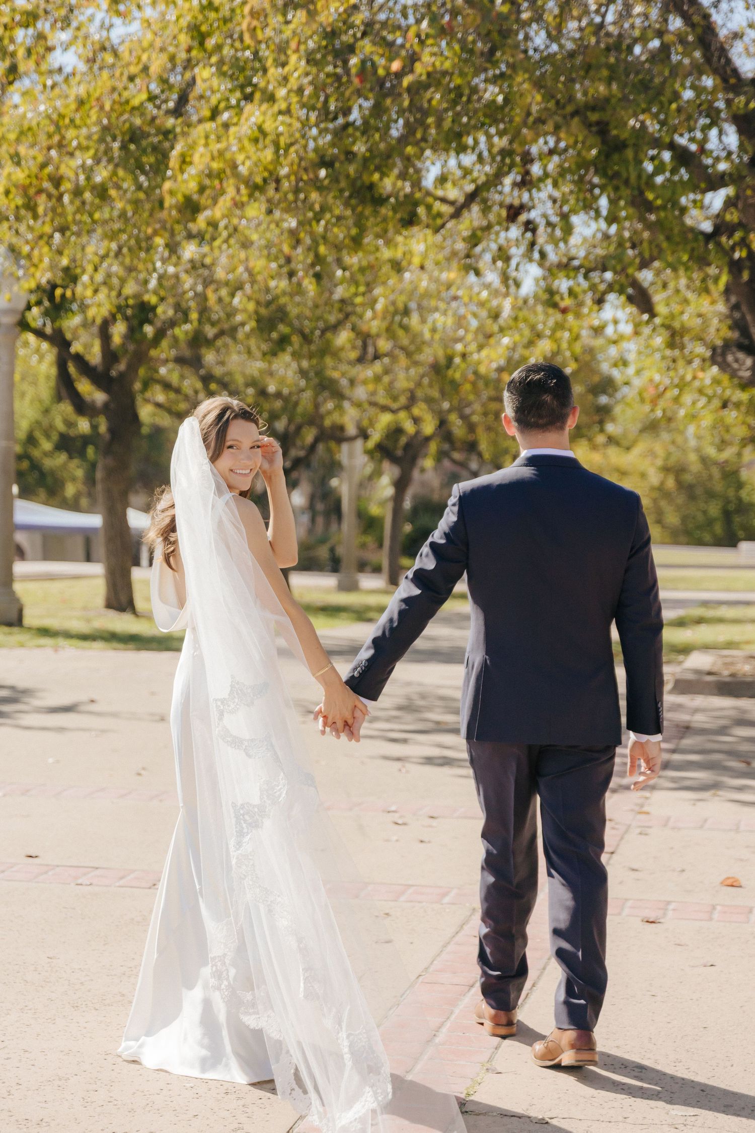 A couple in wedding attire walks together on a tree-lined path during an autumn day.