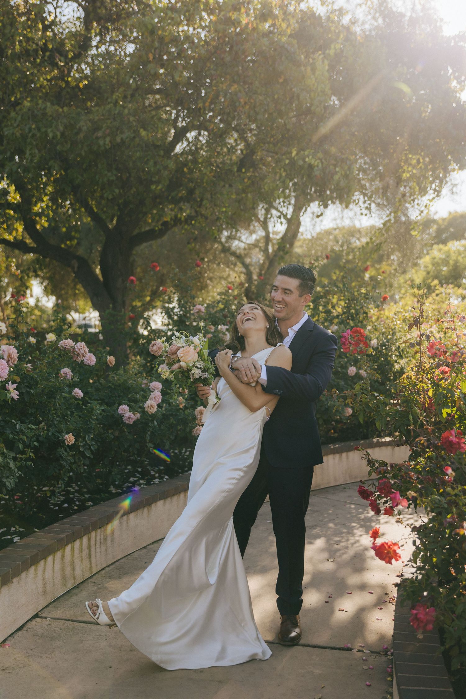 A romantic wedding moment in a garden with blooming roses and sunlit trees as a couple dances together.