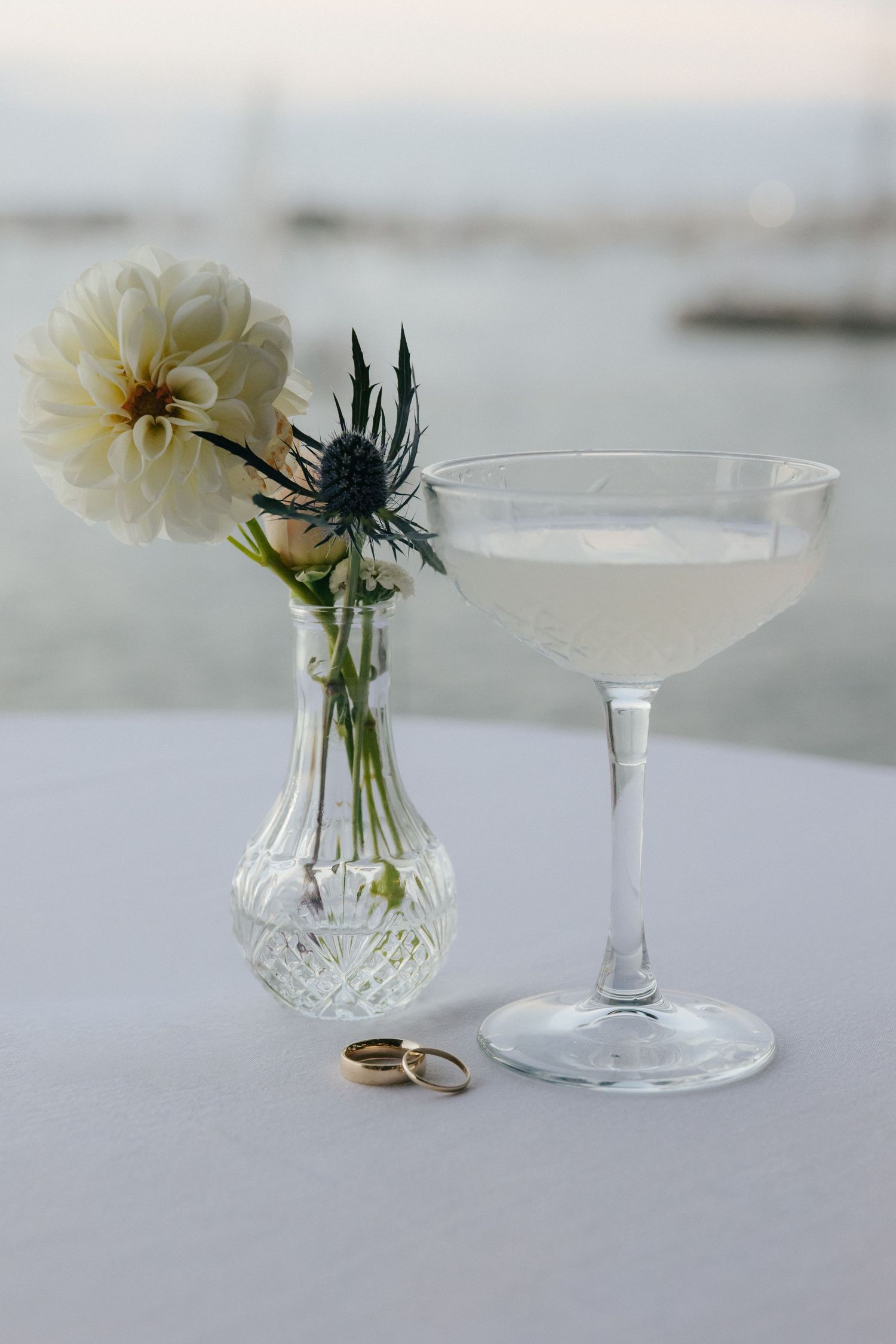 Small white flower in crystal bud vase with wedding rings on white surface.