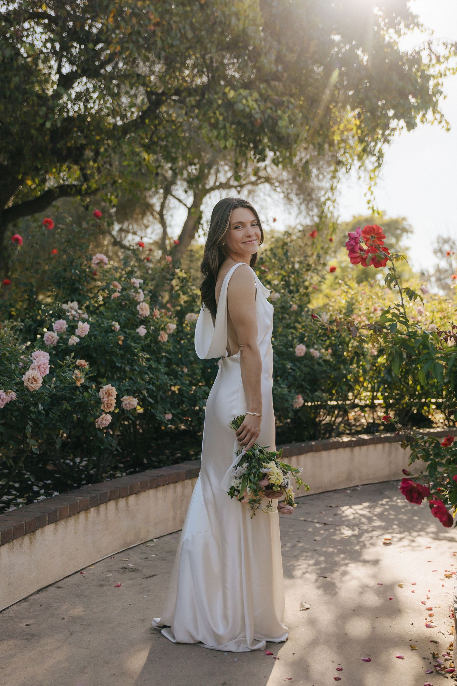 A person in a white wedding dress poses in a garden setting with blooming roses and sunlit trees in the background.