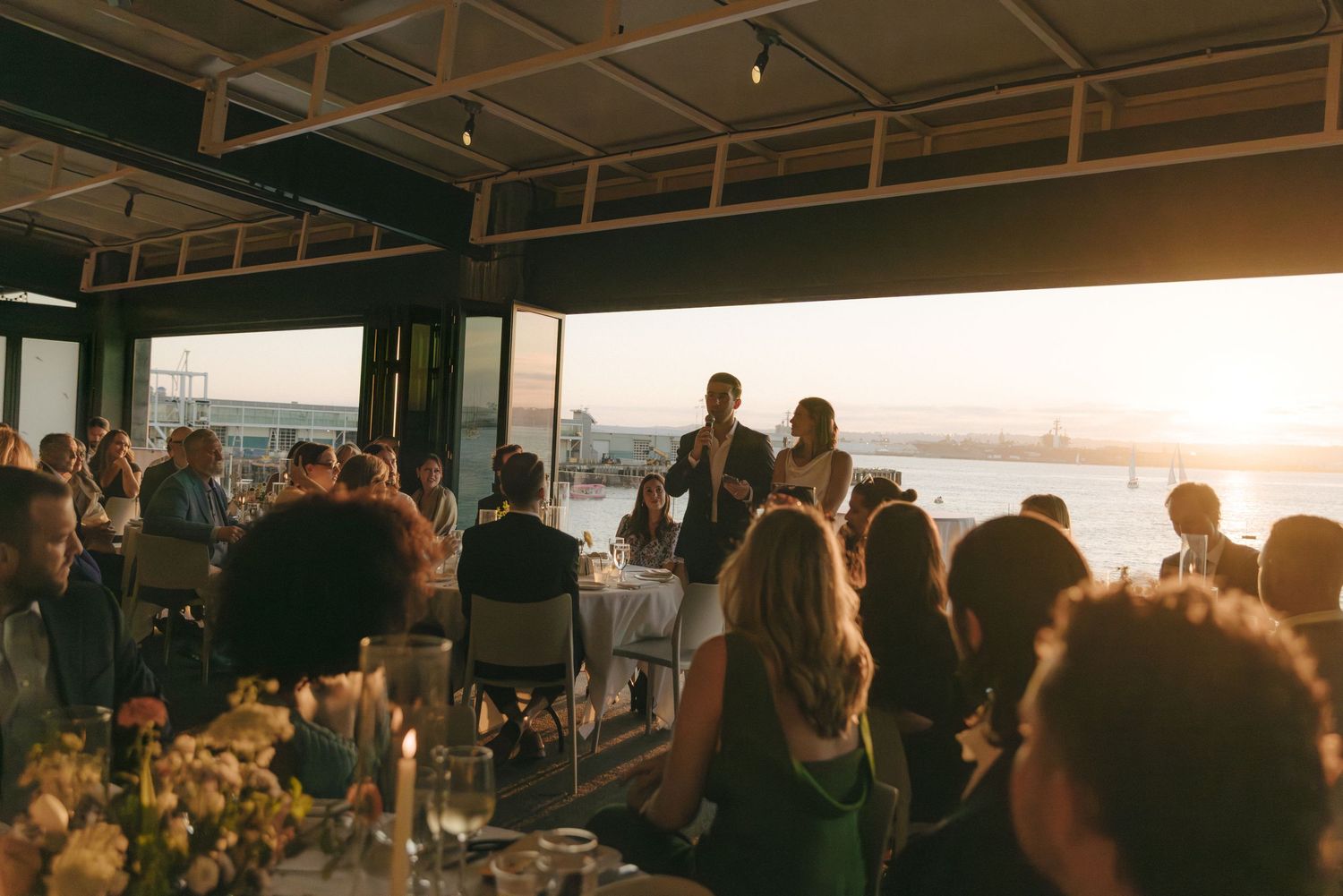 People gather at a waterfront restaurant during golden hour sunset with cityscape views in the background.