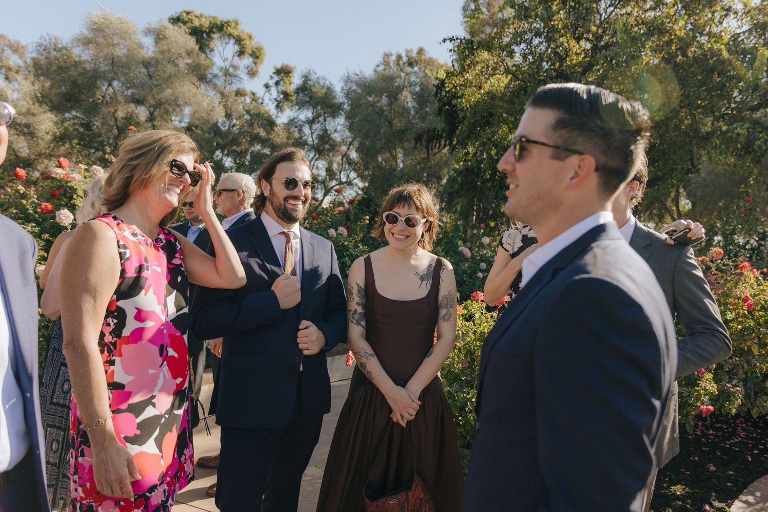A group mingles at an outdoor garden party with guests dressed in formal attire among blooming flowers and trees.
