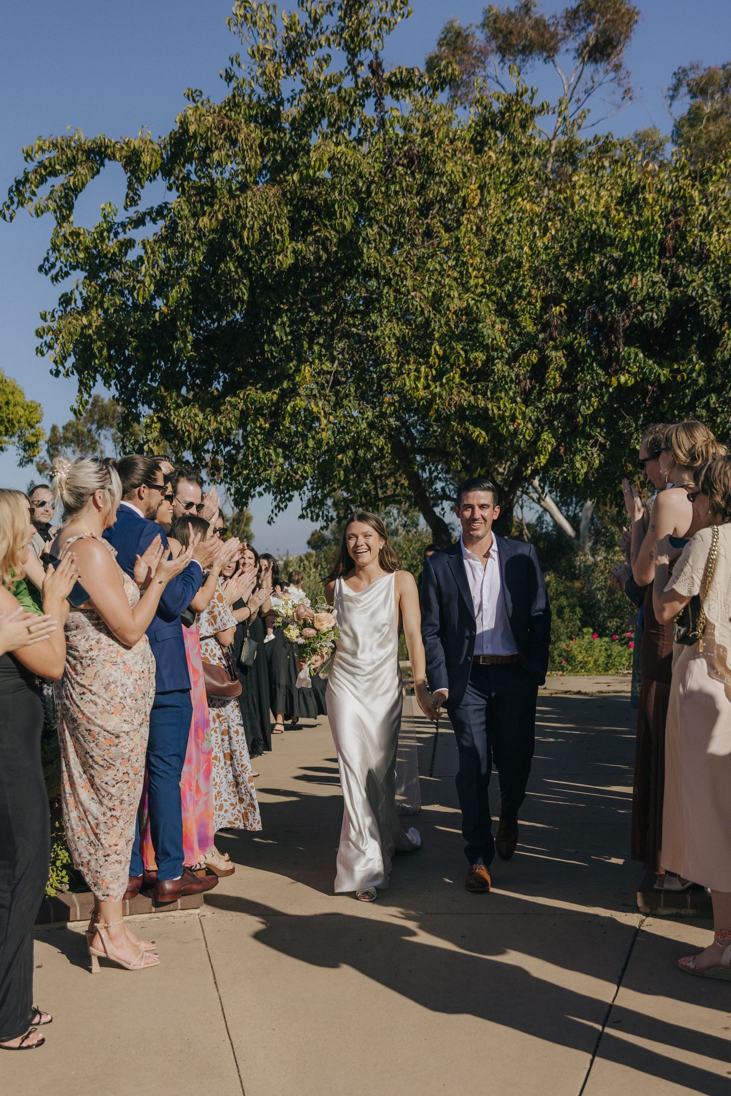 Wedding guests line a pathway outdoors under a leafy tree as a couple walks through in formal attire on a sunny day.