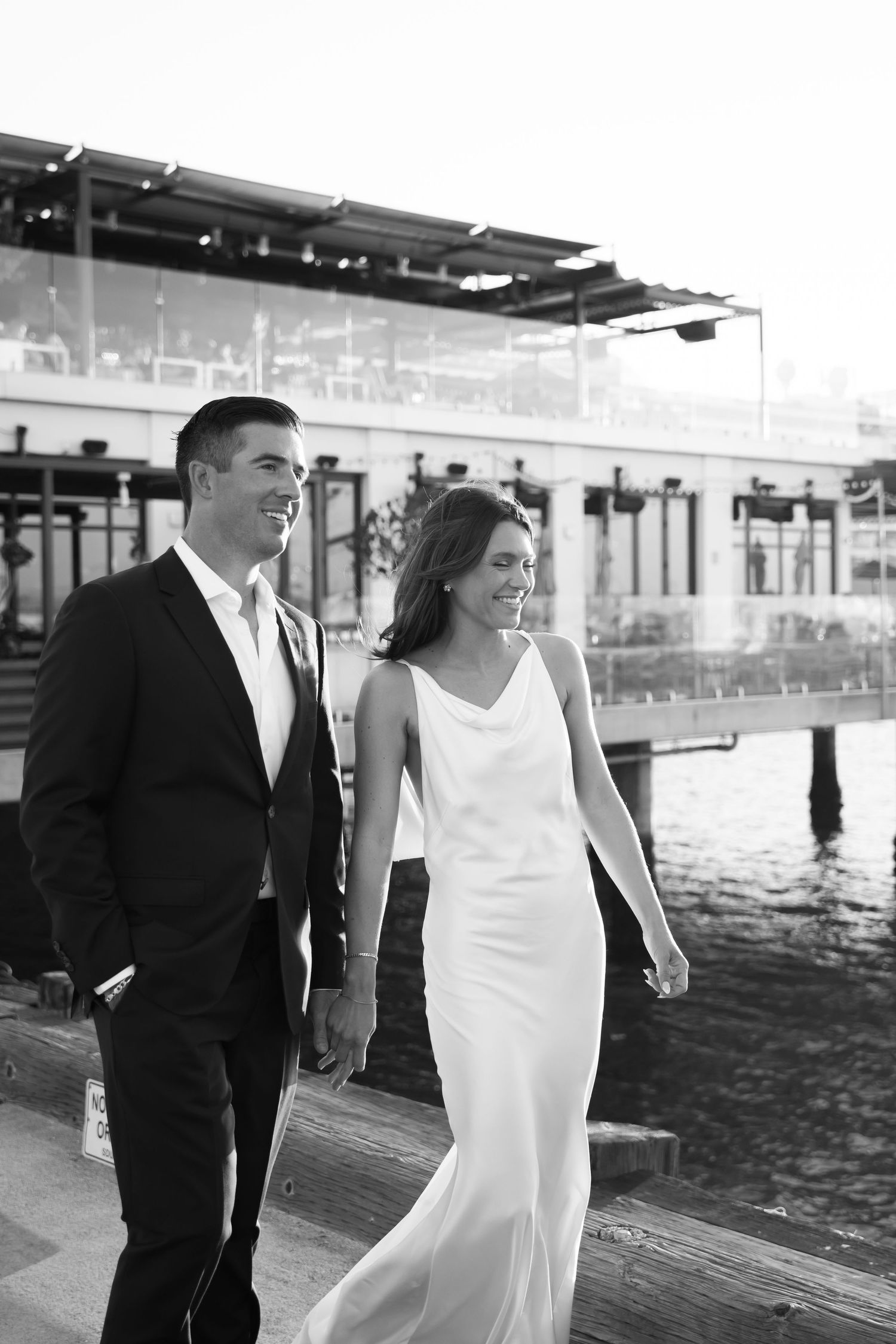A bride and groom walk together on a wooden dock near the water in black and white photography.