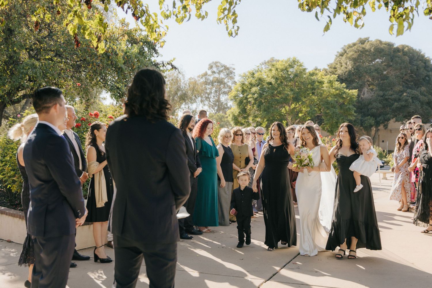 Wedding ceremony guests gather outdoors on a sunny day with trees and greenery in the background.